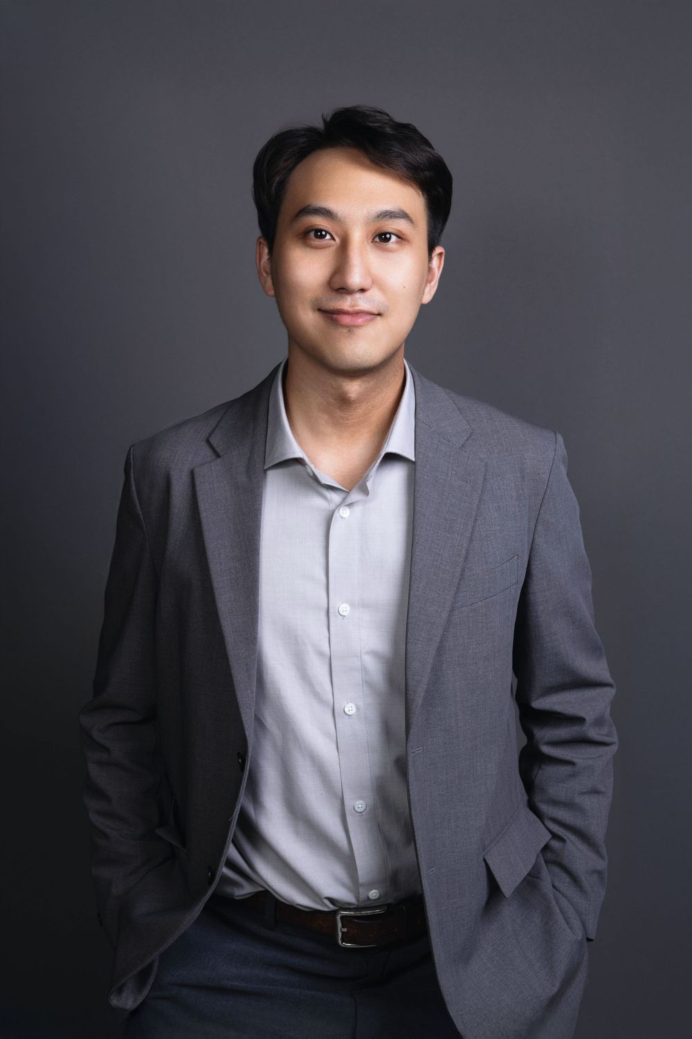 Asian professional man in gray blazer smiling with hands in pockets, corporate headshot on gray background.