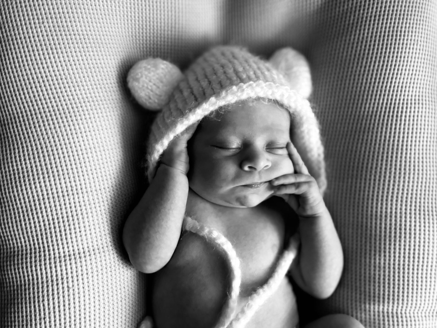 A sleeping baby wearing a knit bear hat lies on a textured surface in black and white.