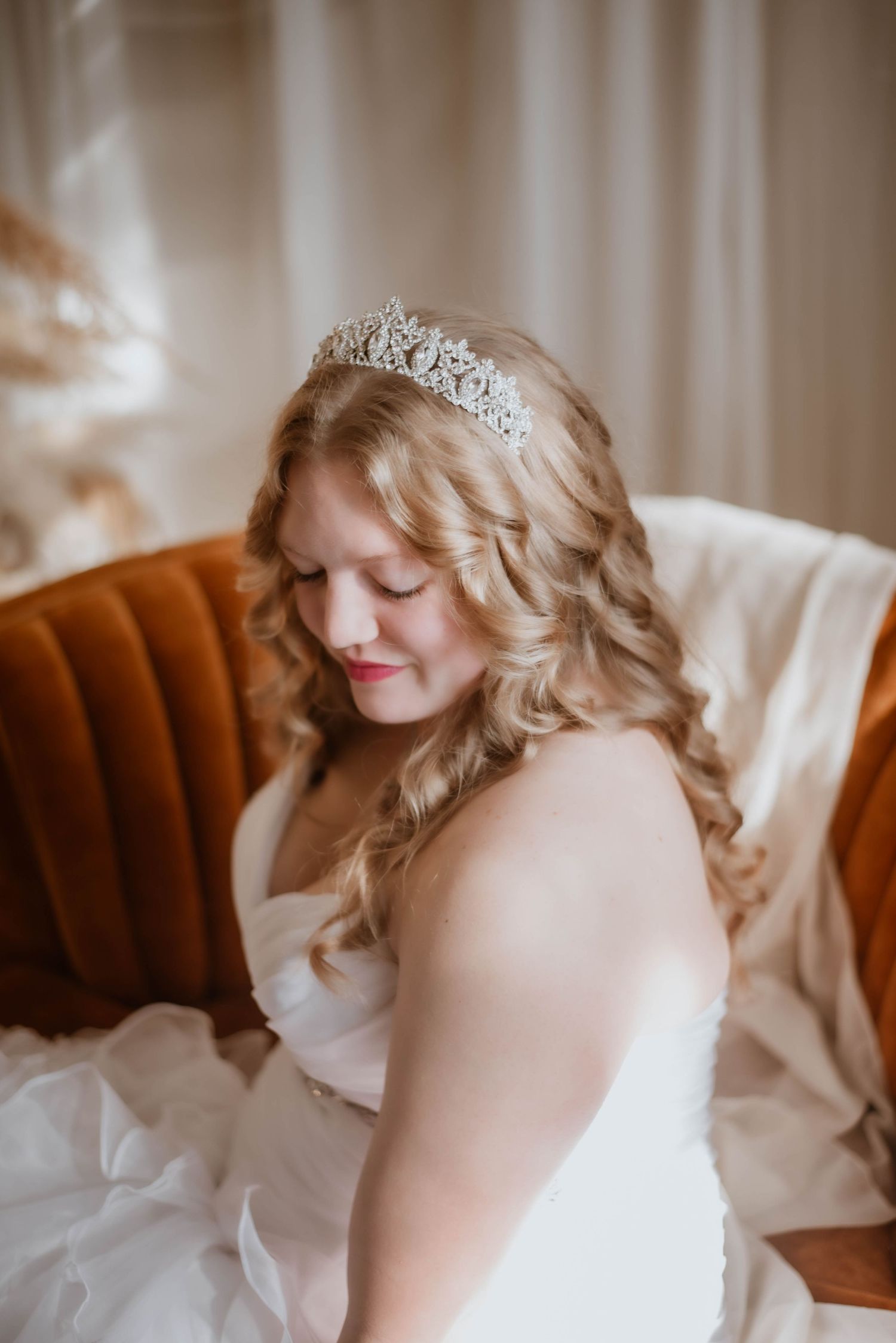Blonde bride wearing a sparkling tiara and white gown seated in a vintage chair.