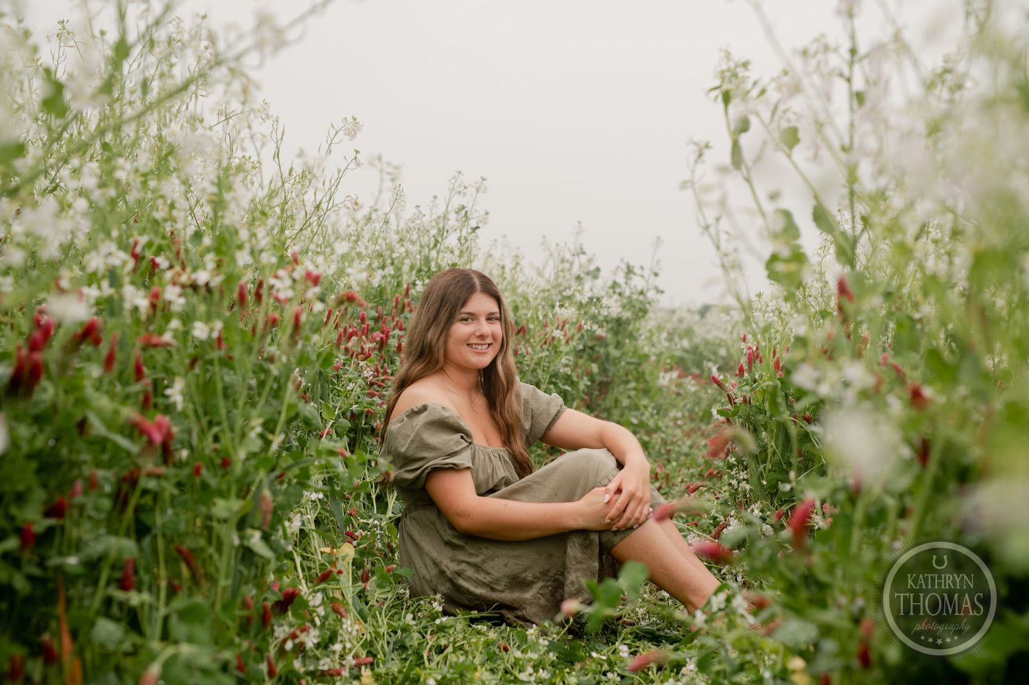 A figure sits casually in a field of wildflowers and tall grass during a dreamy outdoor photo session.