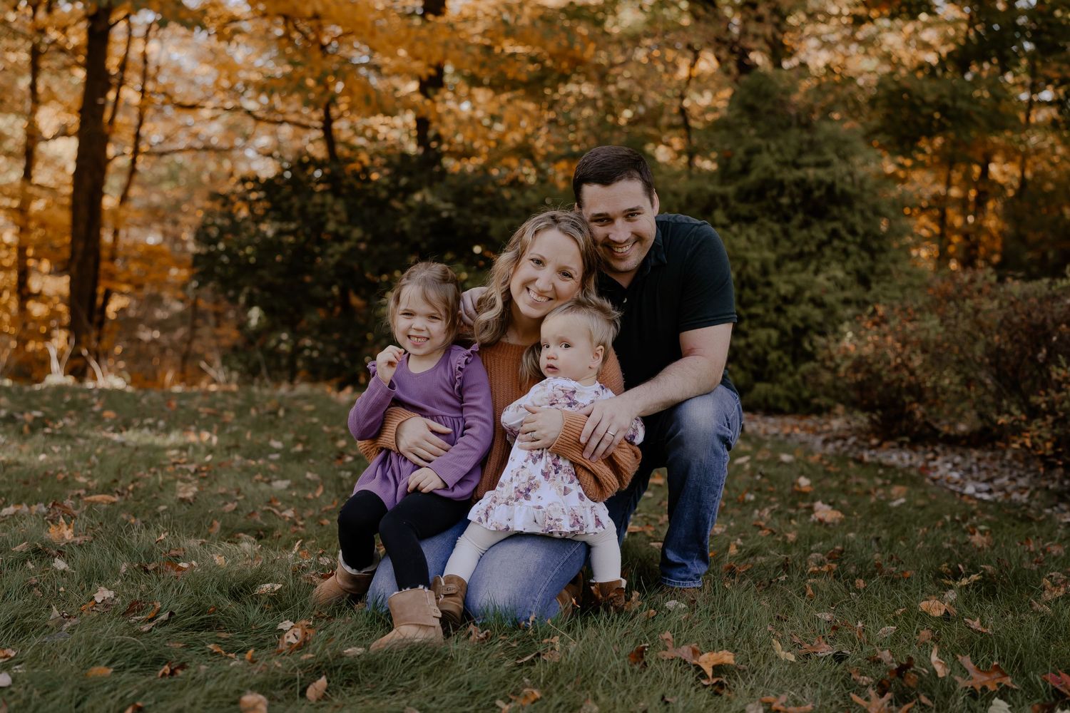 A family sits together on the grass during an autumn sunset with golden fall foliage in the background.