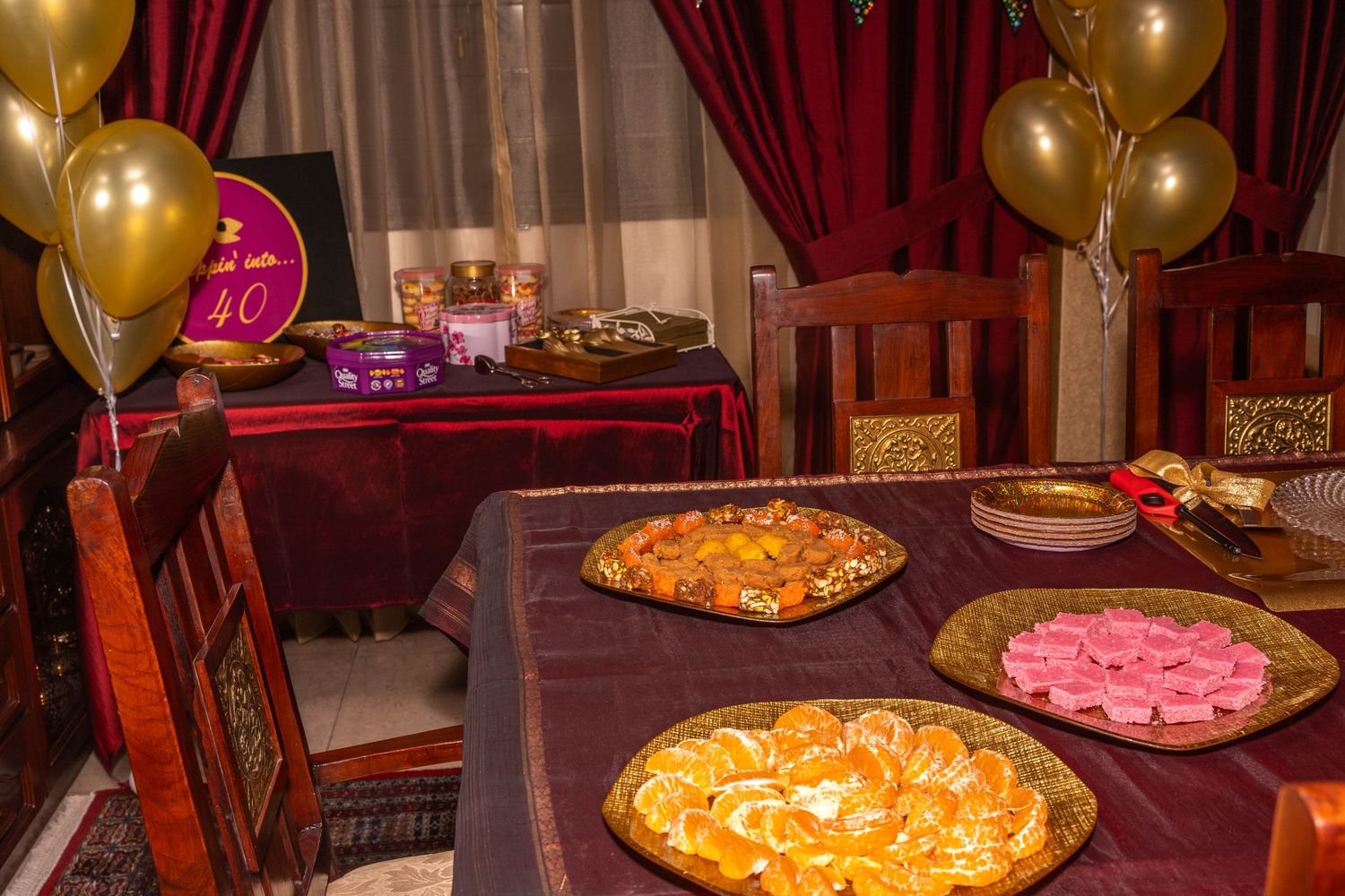 Elegant party table with gold balloons and multiple dishes on burgundy tablecloth.