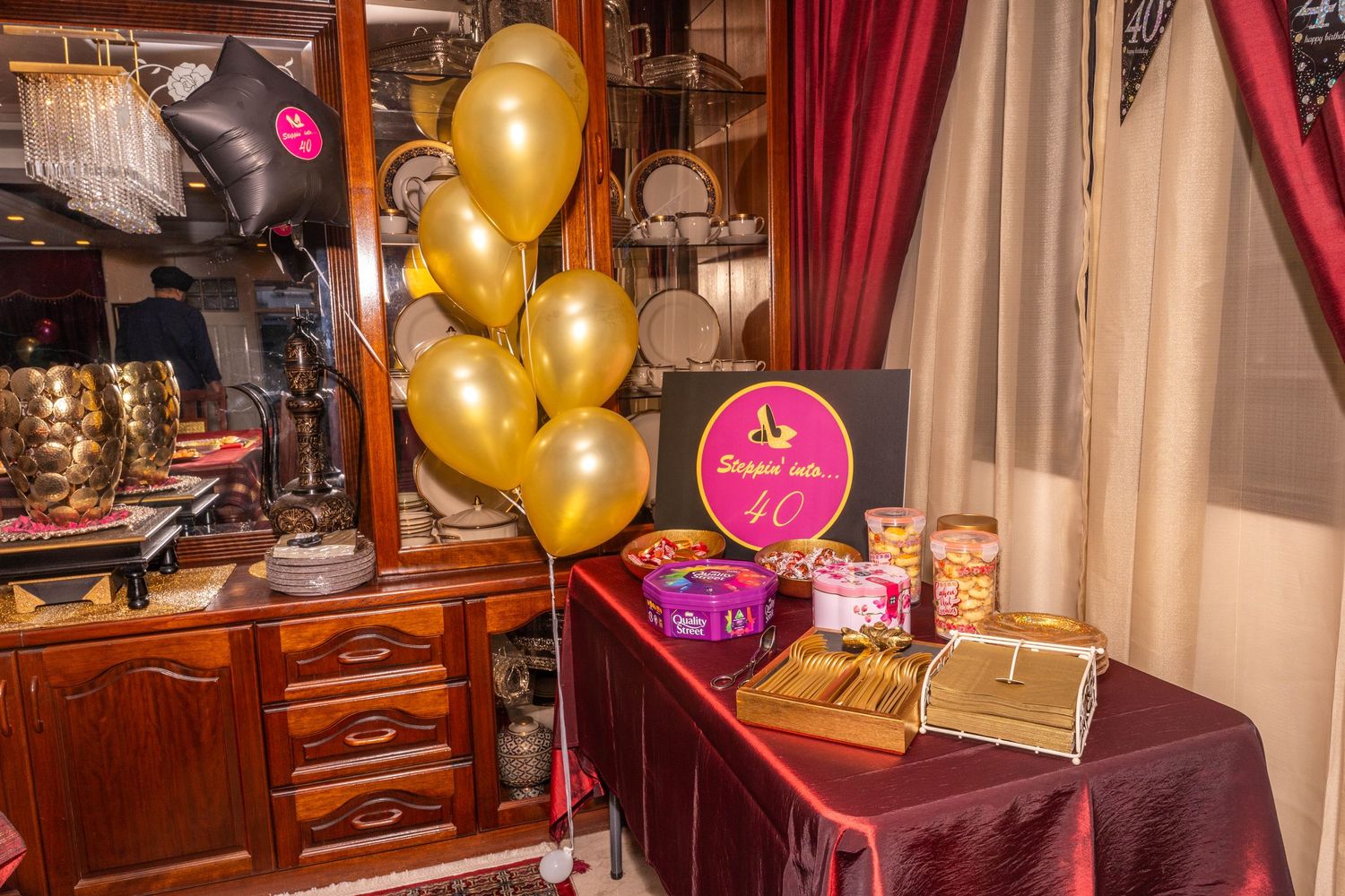 Party setup with gold balloons and pink signage on wooden display cabinet.