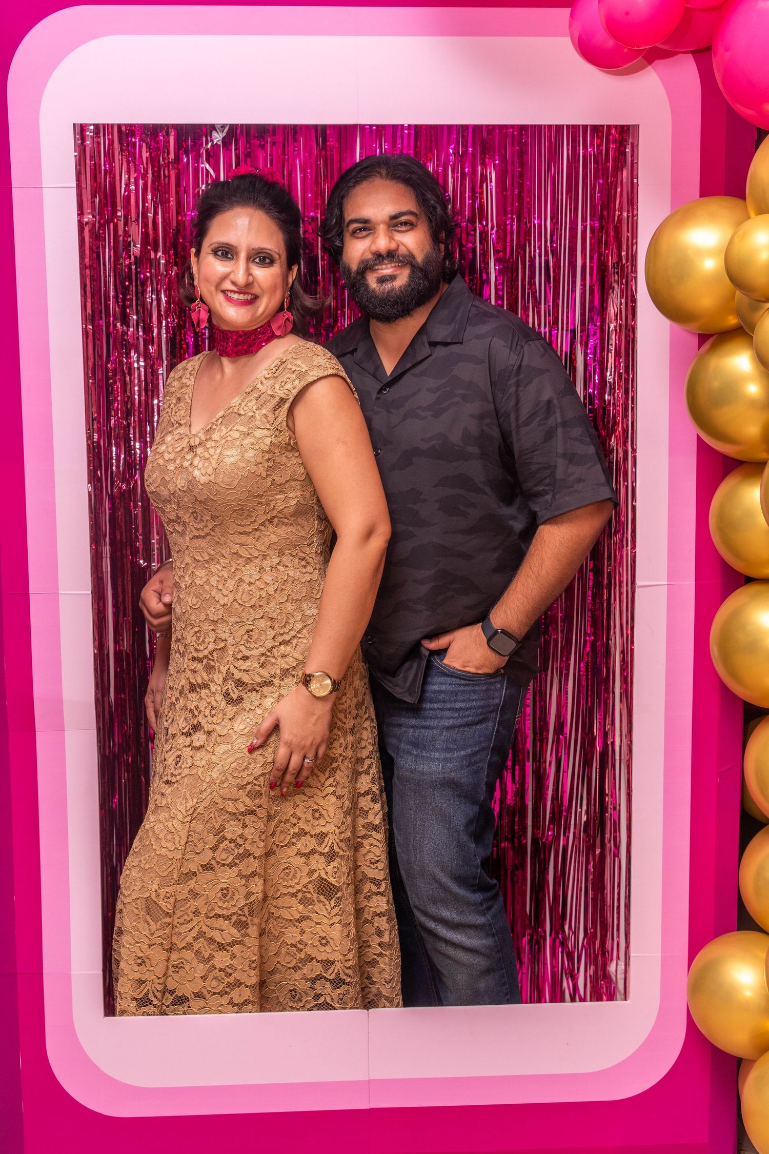 Couple poses against pink metallic backdrop with gold balloon decorations at celebration event.