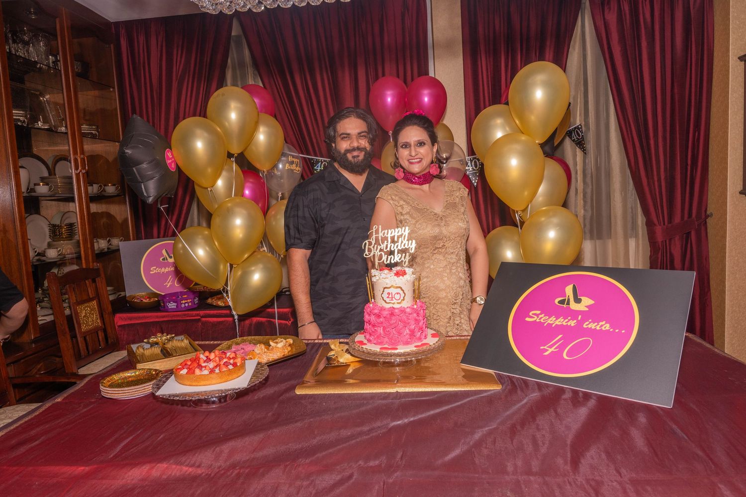Three people posing behind a pink birthday cake with gold and pink balloon decorations.