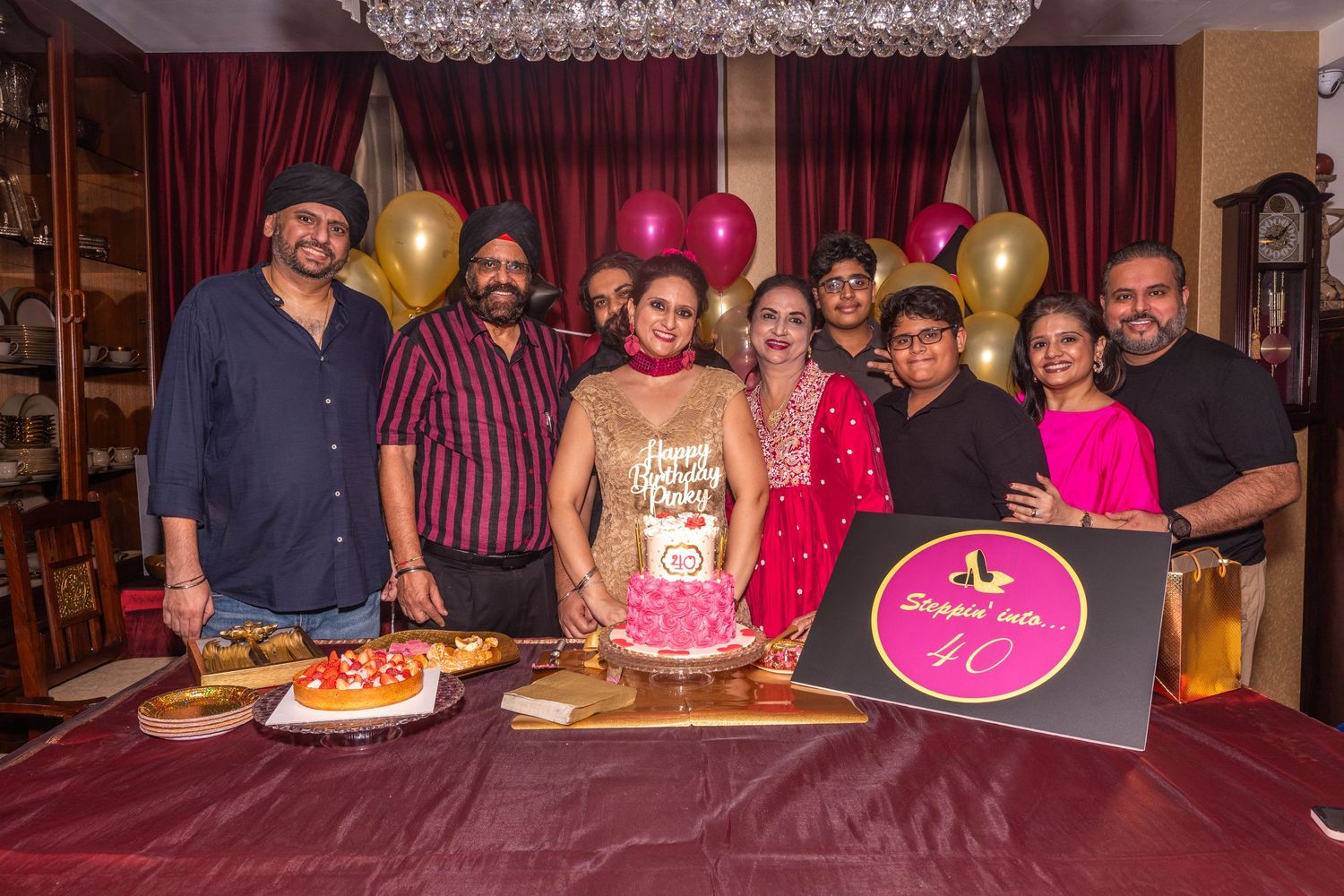 Group photo of party guests gathered around a pink birthday cake with balloons and decorations.
