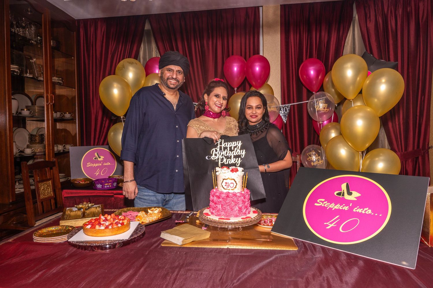 Four people posing with a pink birthday cake surrounded by gold and pink balloons.