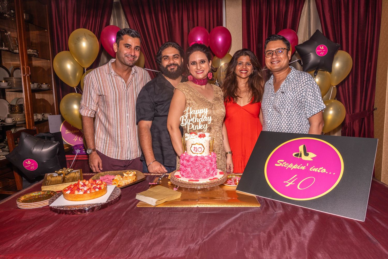 Five guests posing at a birthday celebration with cake and festive balloon decorations.