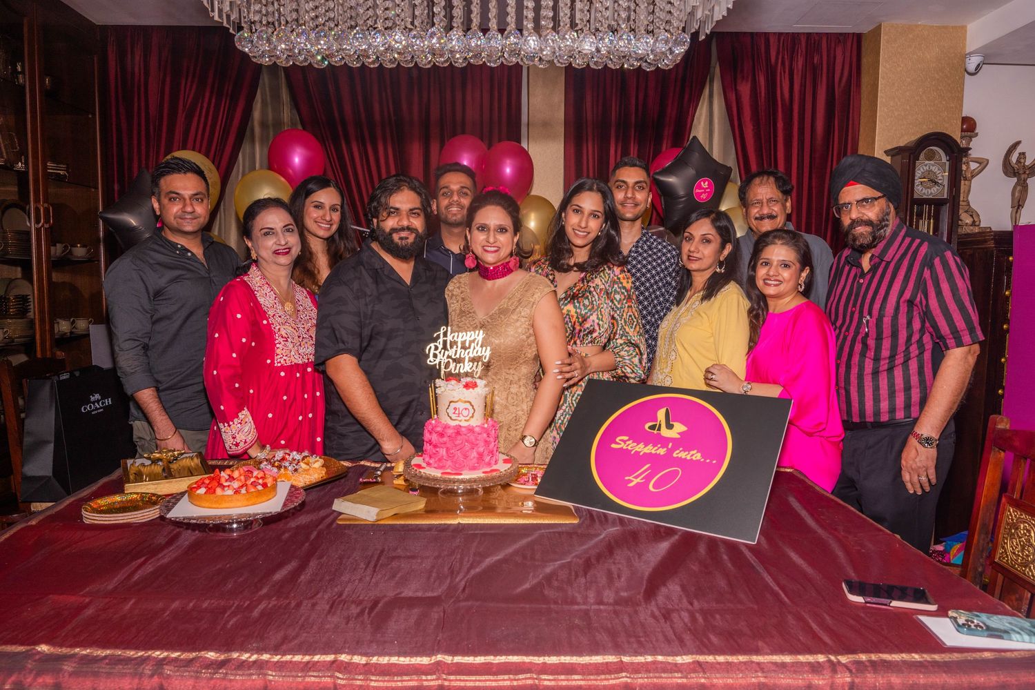 A festive birthday celebration with a group gathered around a cake and decorative balloons under a chandelier.