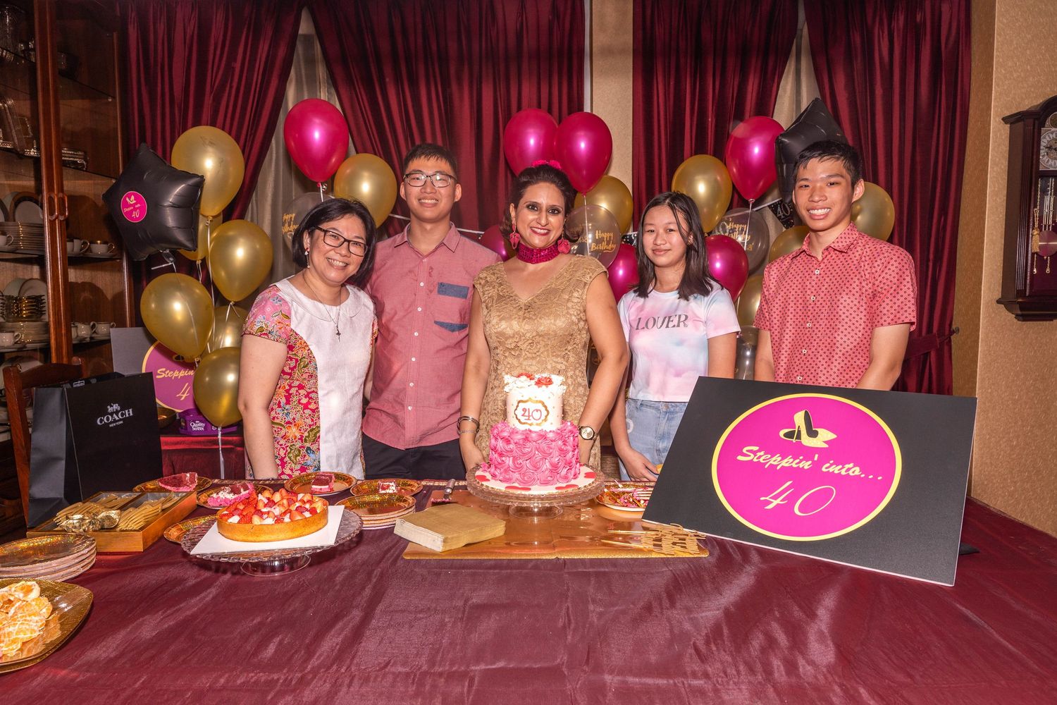 Birthday party gathering with pink and gold balloons, decorated cake, and a festive sign on burgundy tablecloth.