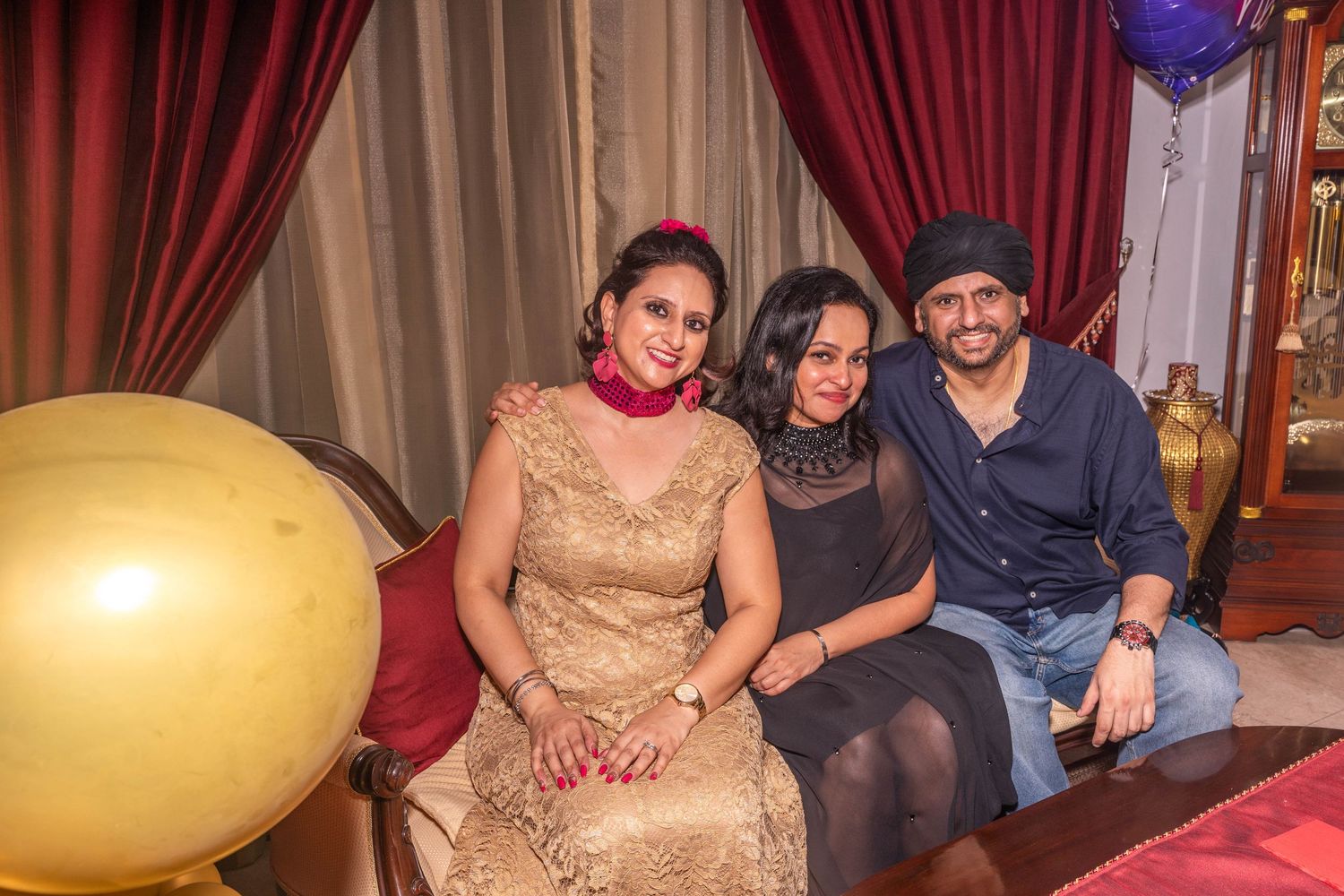 Three people sitting on a couch at a party with gold balloons in the background.