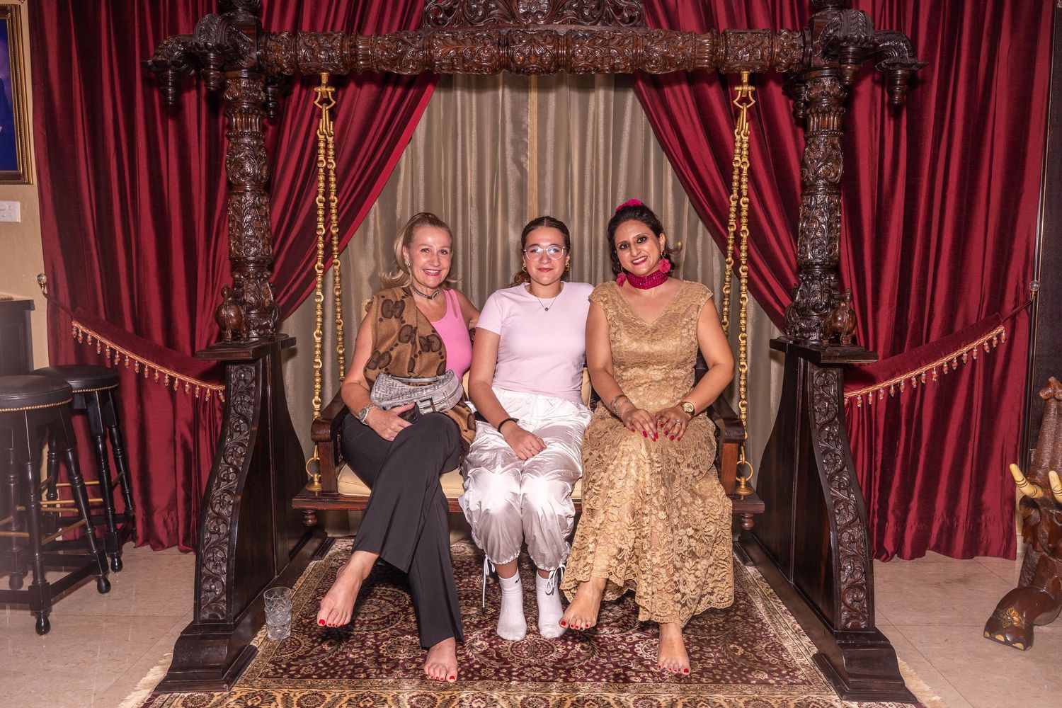 Three people seated in an ornate wooden throne-like structure with red velvet curtains.