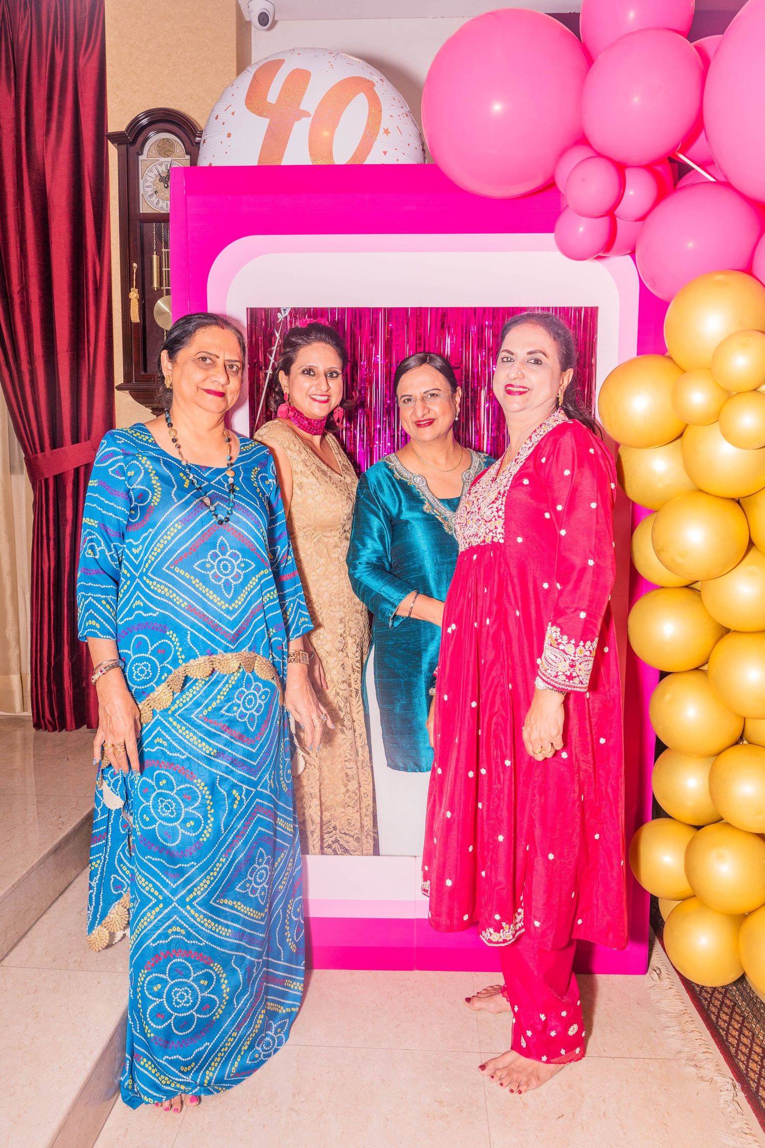 Four people in colorful traditional attire posing by a pink photo frame with gold balloon decorations.