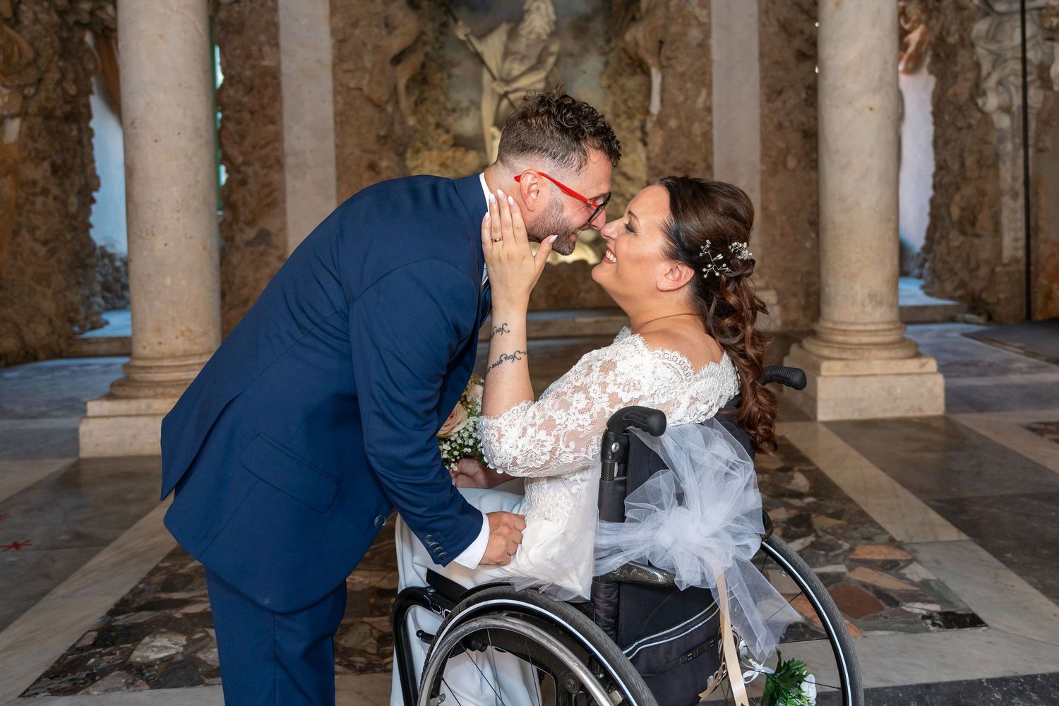 Radiant bride in wheelchair smiling with wedding bouquet on stone steps.