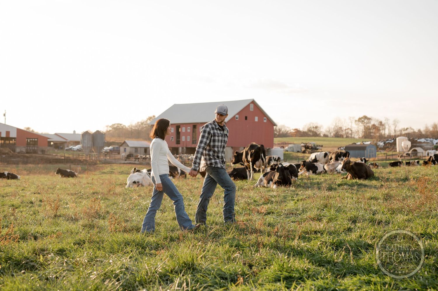 Two people walk hand in hand across their farm pasture with cattle and a red barn in view.