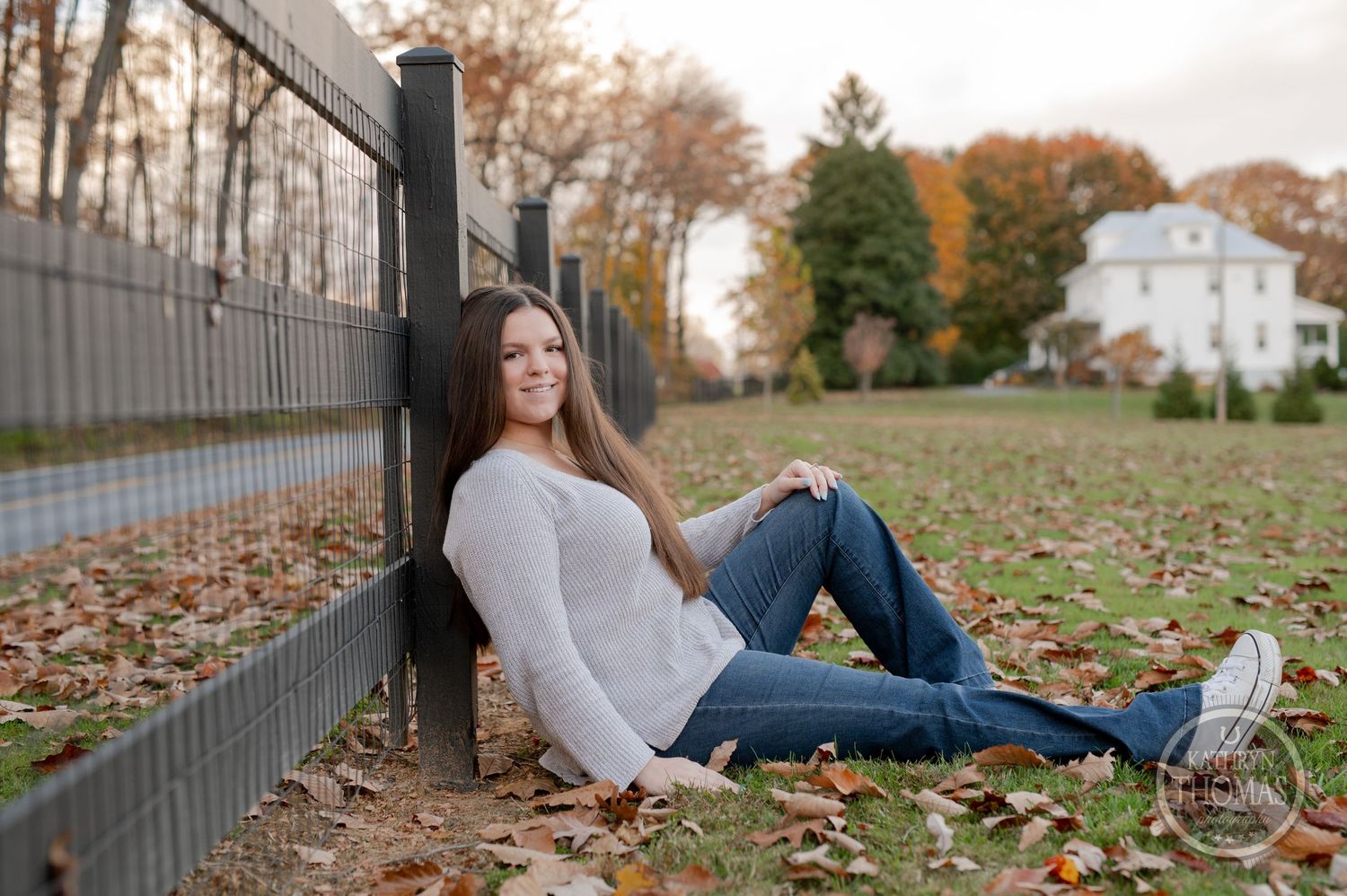 A person in a light sweater and jeans sits against a black fence surrounded by autumn leaves on the ground.