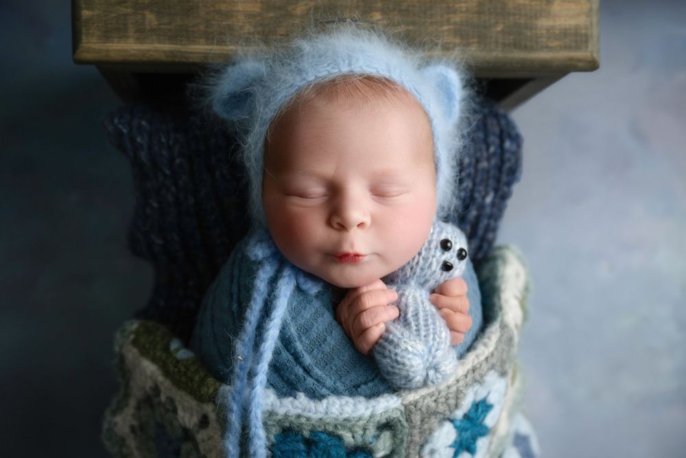 Newborn baby wrapped in blue knit with fuzzy bear ears against wooden backdrop.