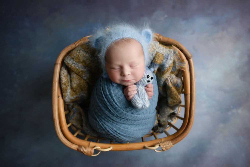 Sleeping newborn in blue wrap lying in wicker basket with floral cushioning.