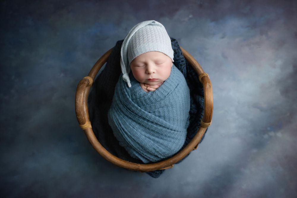 Sleeping newborn in blue wrap and white hat nestled in wooden bowl.