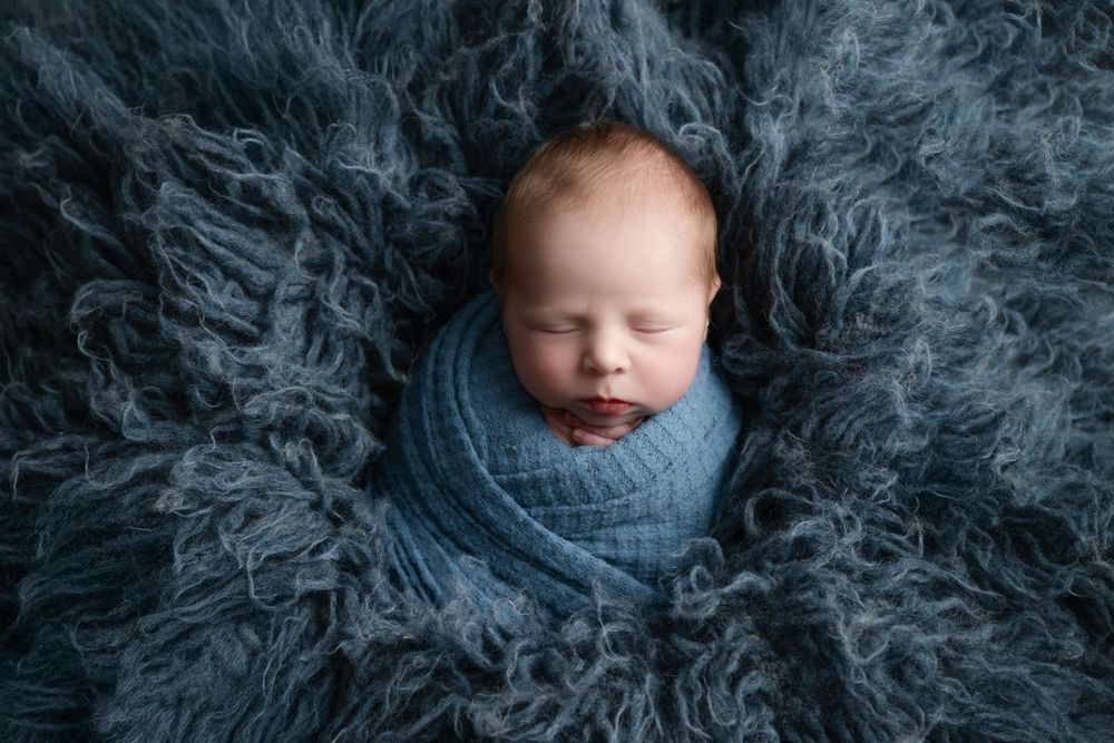 Sleeping newborn wrapped in blue knit against grey fluffy textured backdrop.