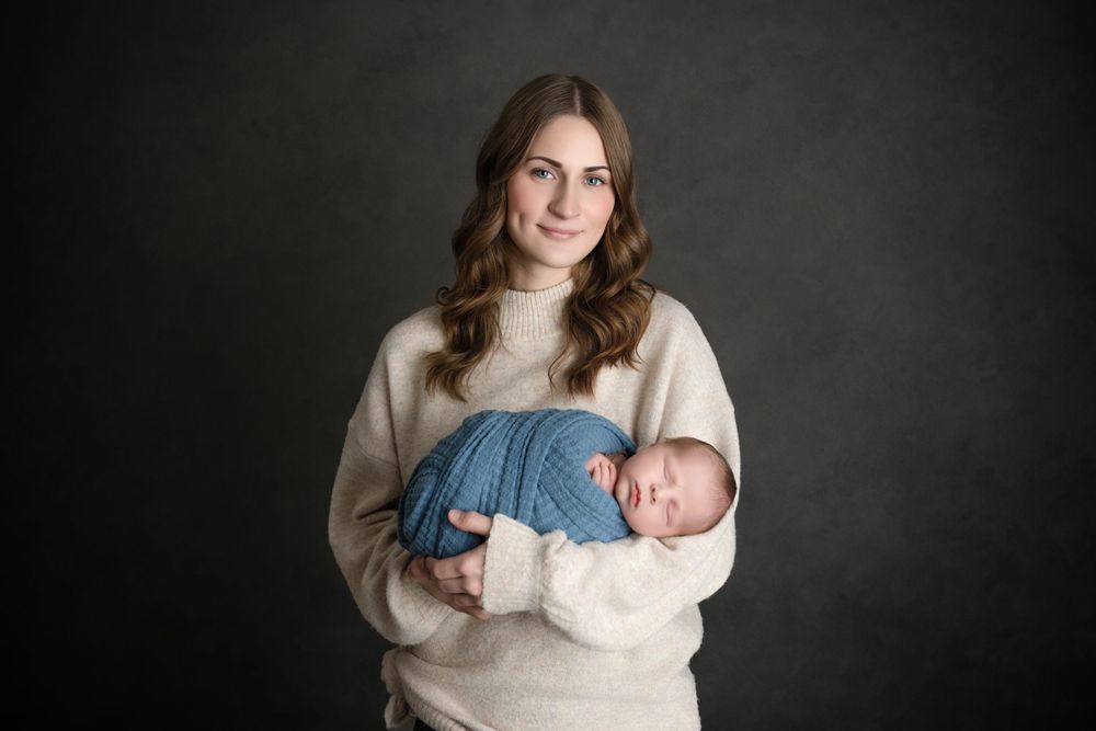 Studio portrait of mother in cream sweater holding newborn wrapped in blue.