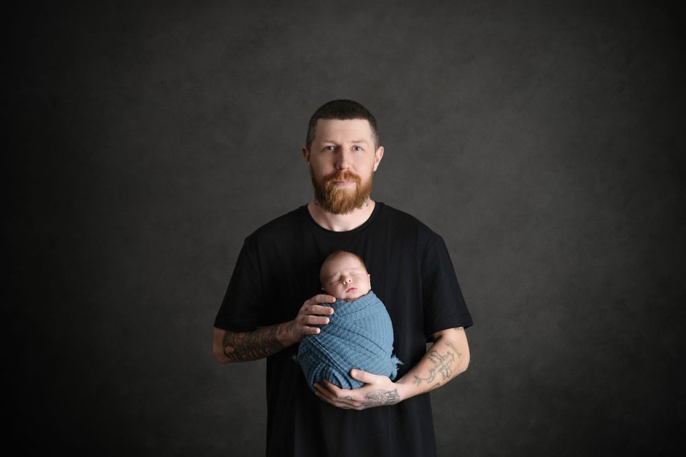 Studio portrait of father in black shirt holding newborn wrapped in blue.