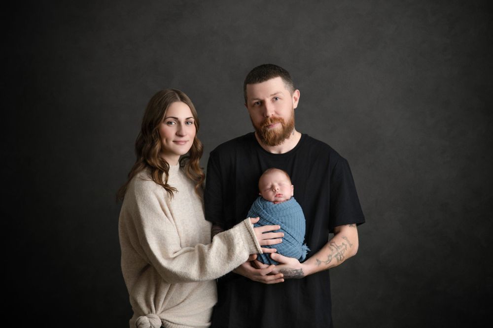 Family portrait featuring parents holding their newborn baby against a dark gray backdrop.