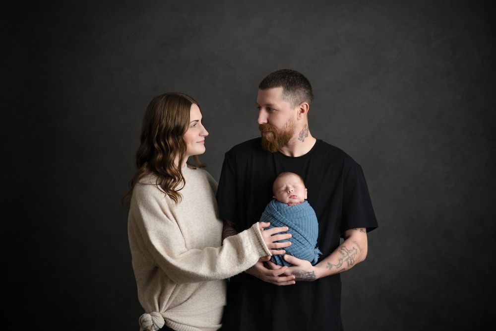 Parents smiling lovingly at their infant while holding them against a dark background.