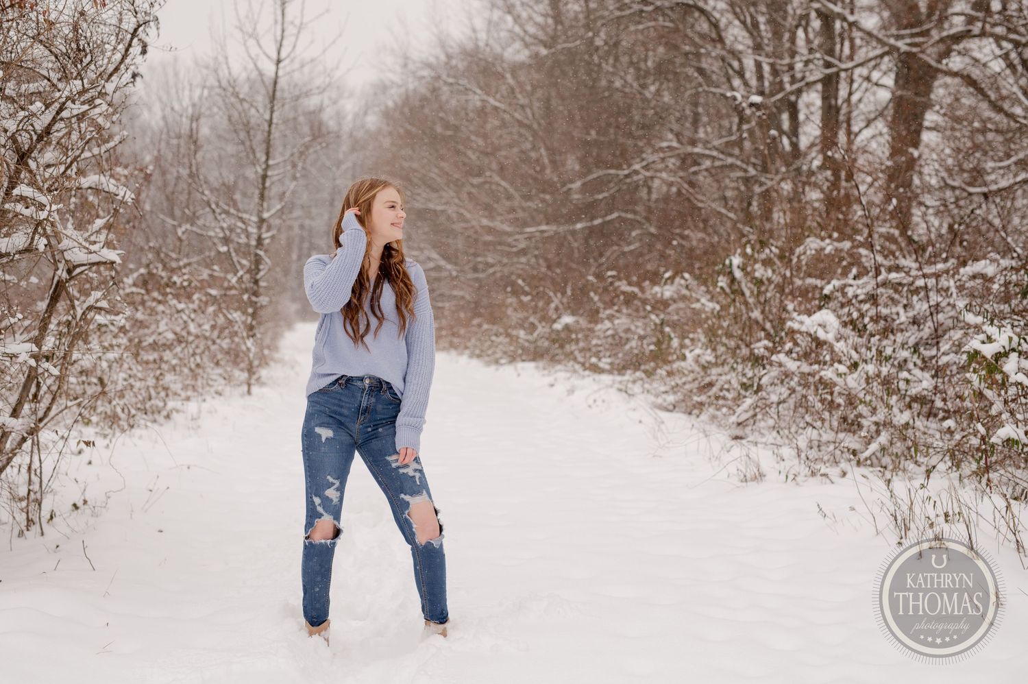 Casual winter outfit photograph with ripped jeans against a snow-covered forest backdrop.