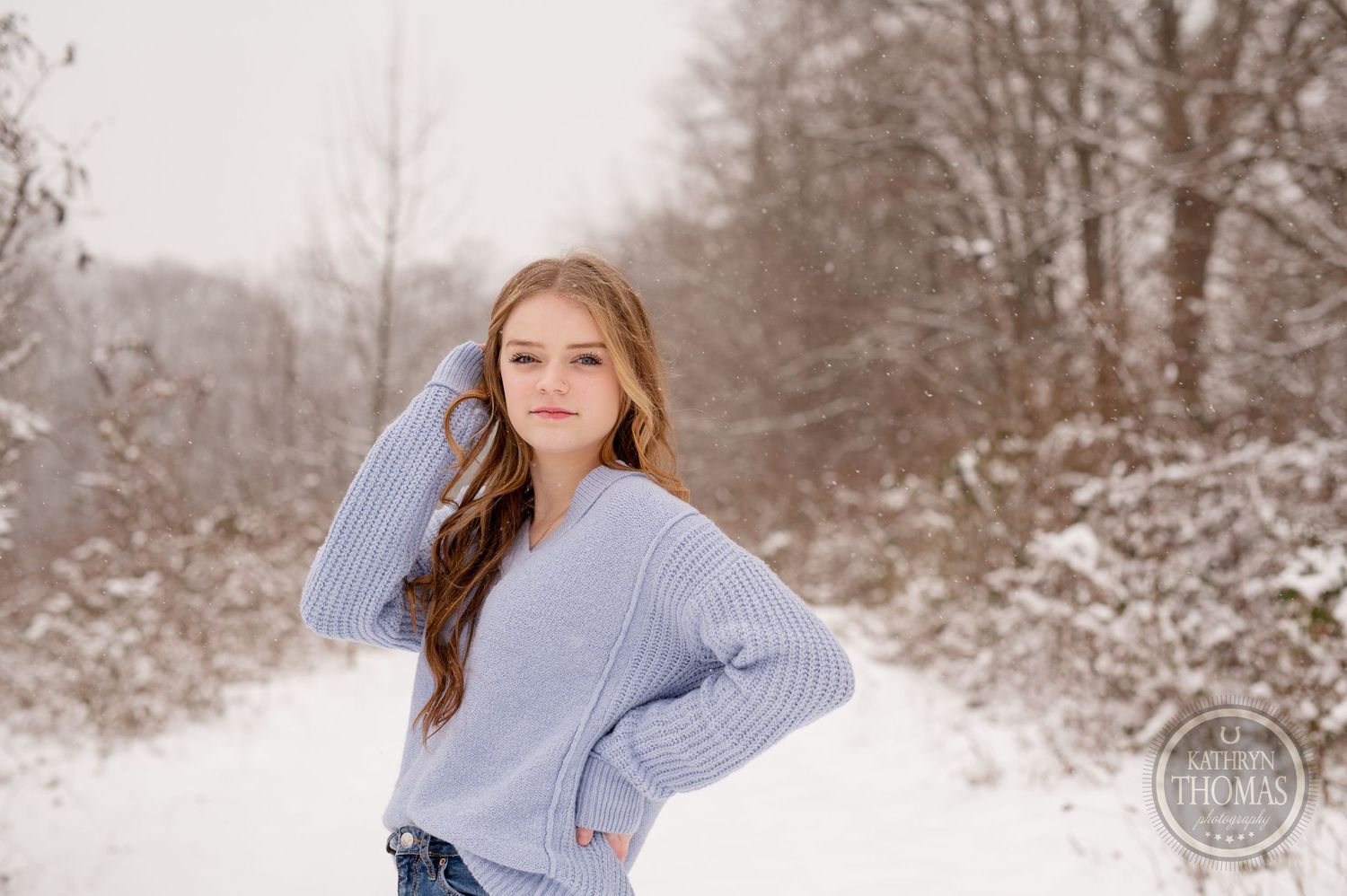 Person in light blue sweater poses casually against winter forest backdrop.