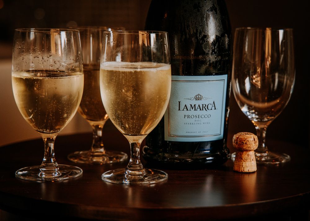 Filled champagne glasses with La Marca Prosecco bottle and cork on wooden surface.