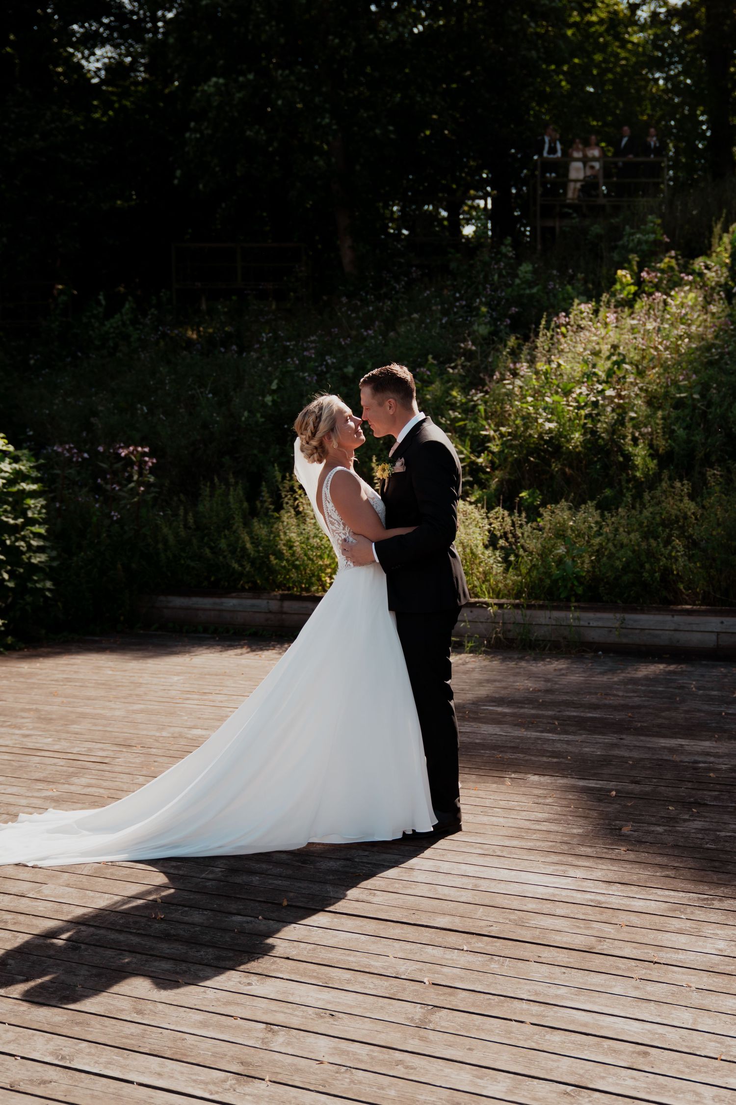 A romantic wedding couple poses on a wooden deck in a garden setting during golden hour.