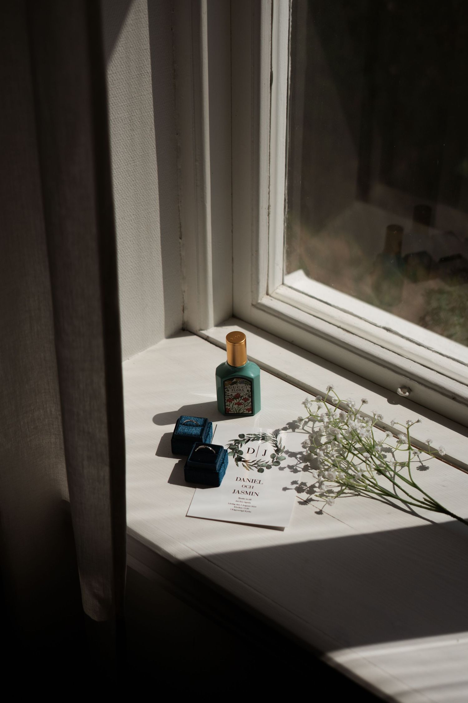 Wedding details arranged on a sunlit windowsill including rings, flowers, and a small blue box.