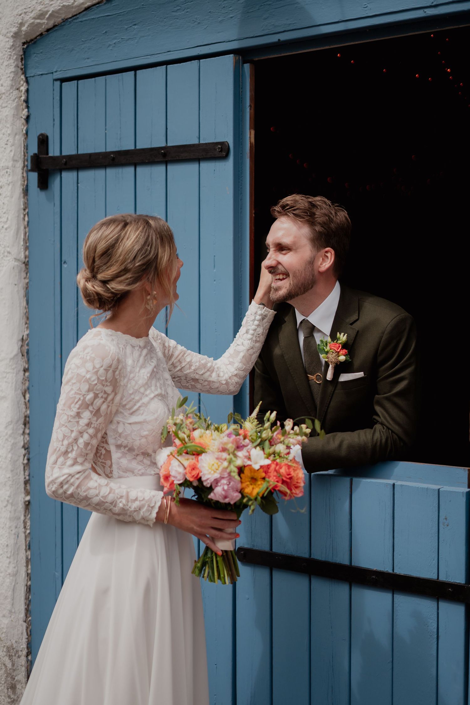 A couple interacts playfully through blue barn doors with colorful flower bouquets.