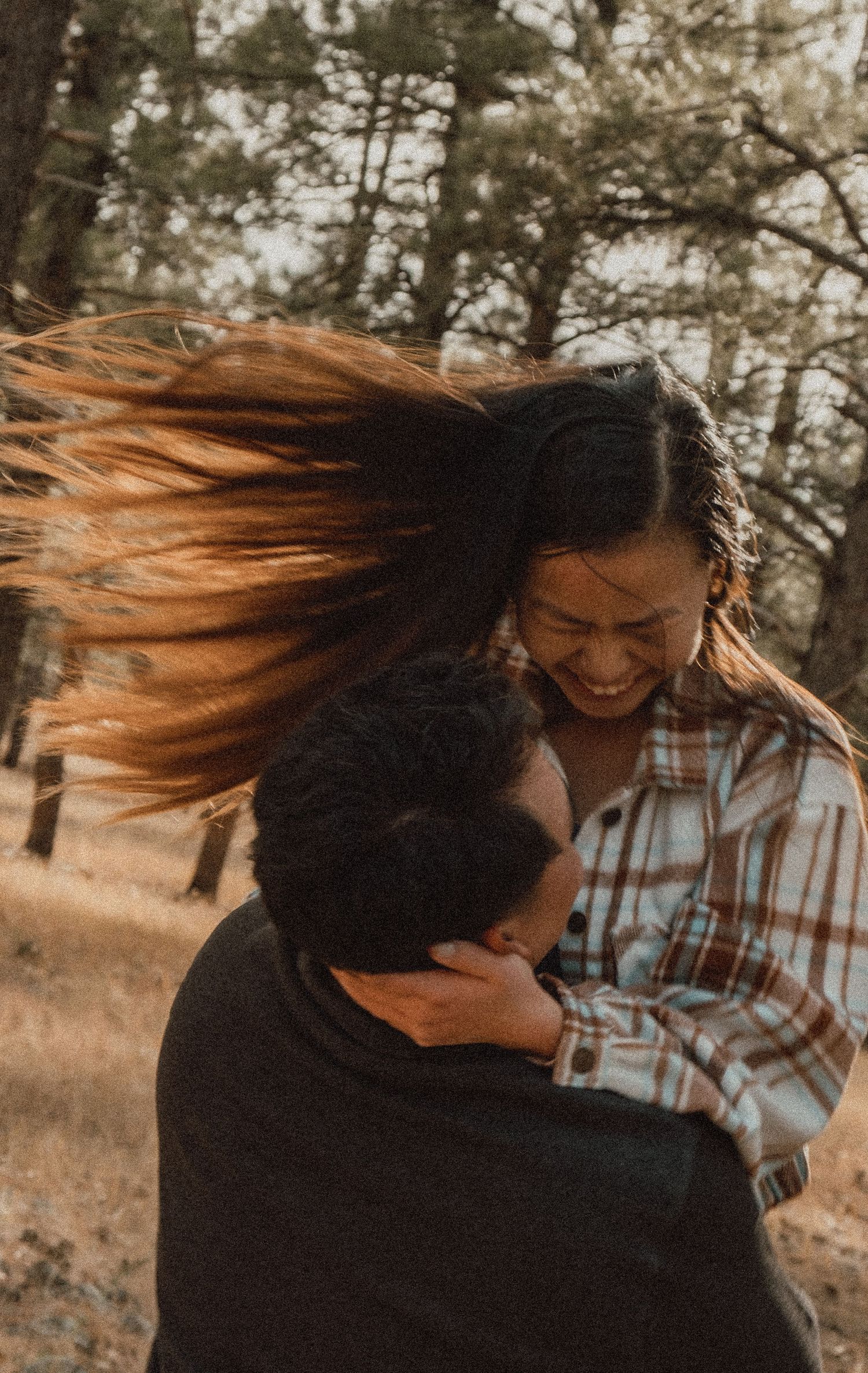 Two people laughing and embracing outdoors while their hair blows in the wind in a dreamy autumn forest setting.