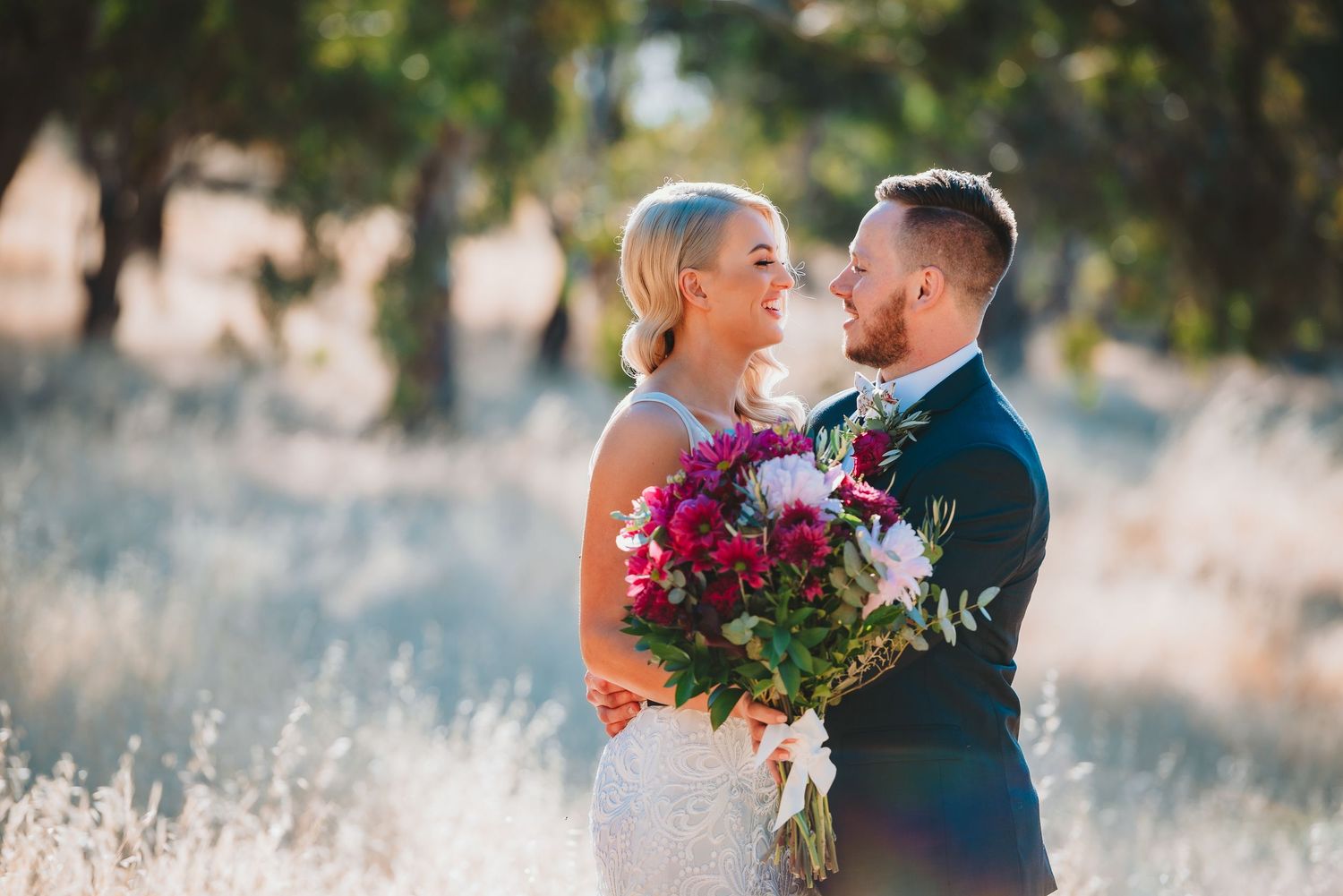 A romantic newlywed couple embracing in a sun-lit meadow with vibrant fuchsia and purple floral bouquet.