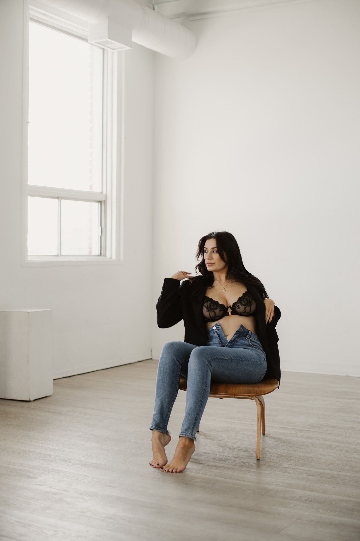 Woman in jeans and dark top sits barefoot on wooden chair by window in minimalist white room.