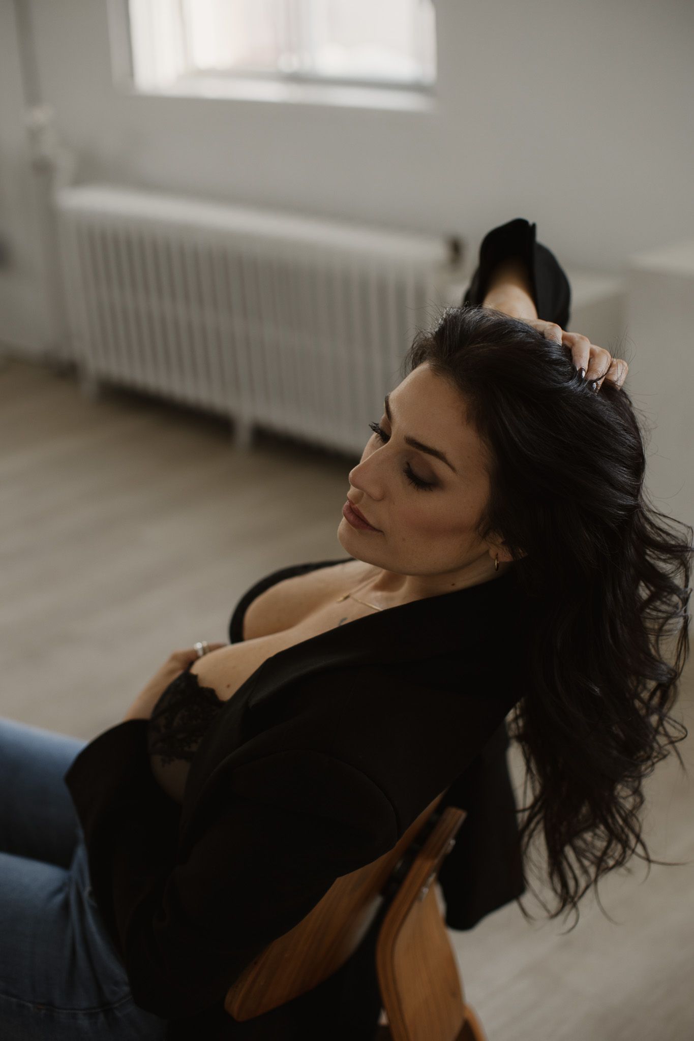 Woman with long dark hair wearing black top and jeans rests on floor near window in moody indoor portrait setting.