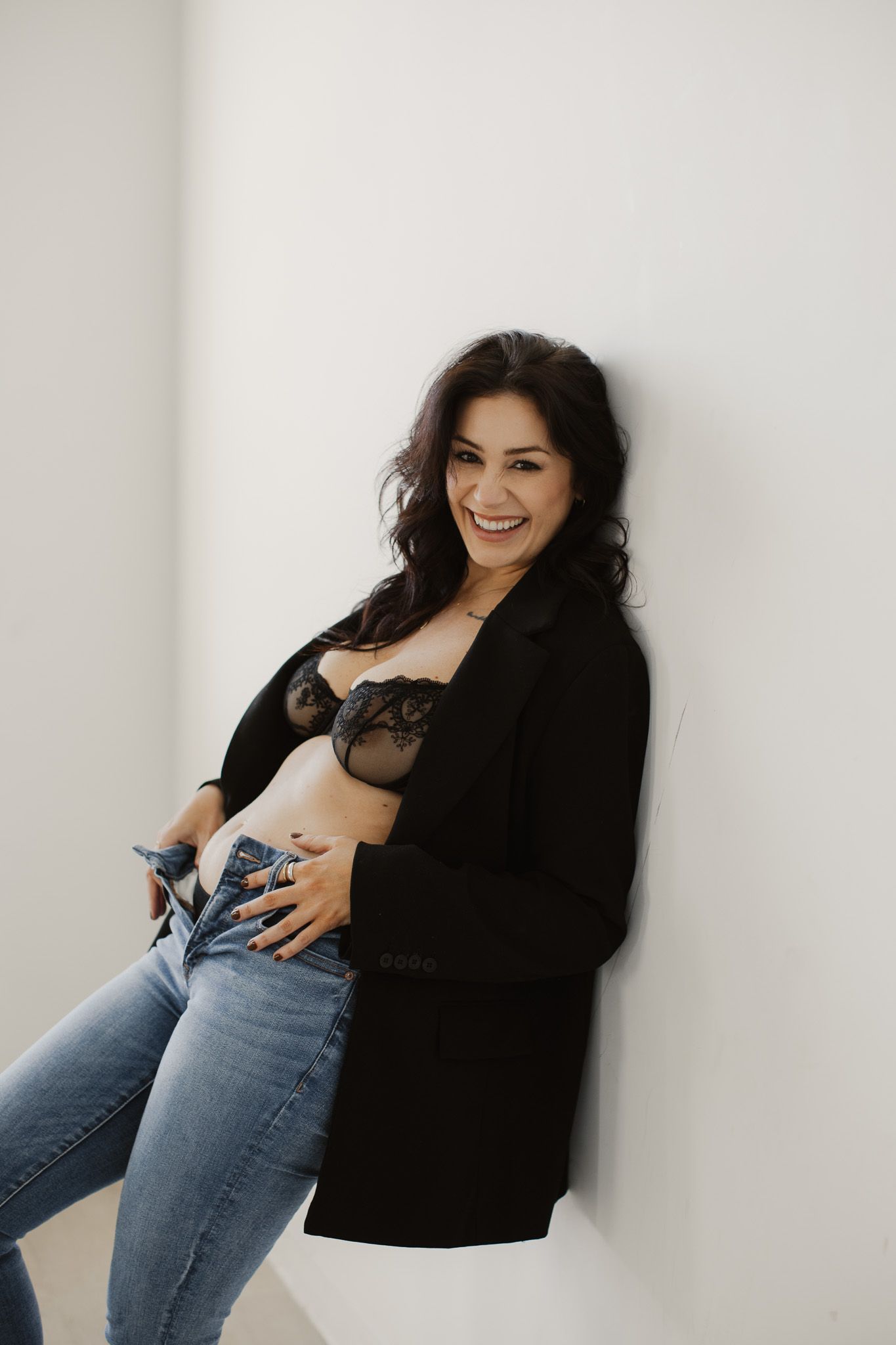 A woman wearing a black cardigan and jeans poses against a white wall with a joyful expression.