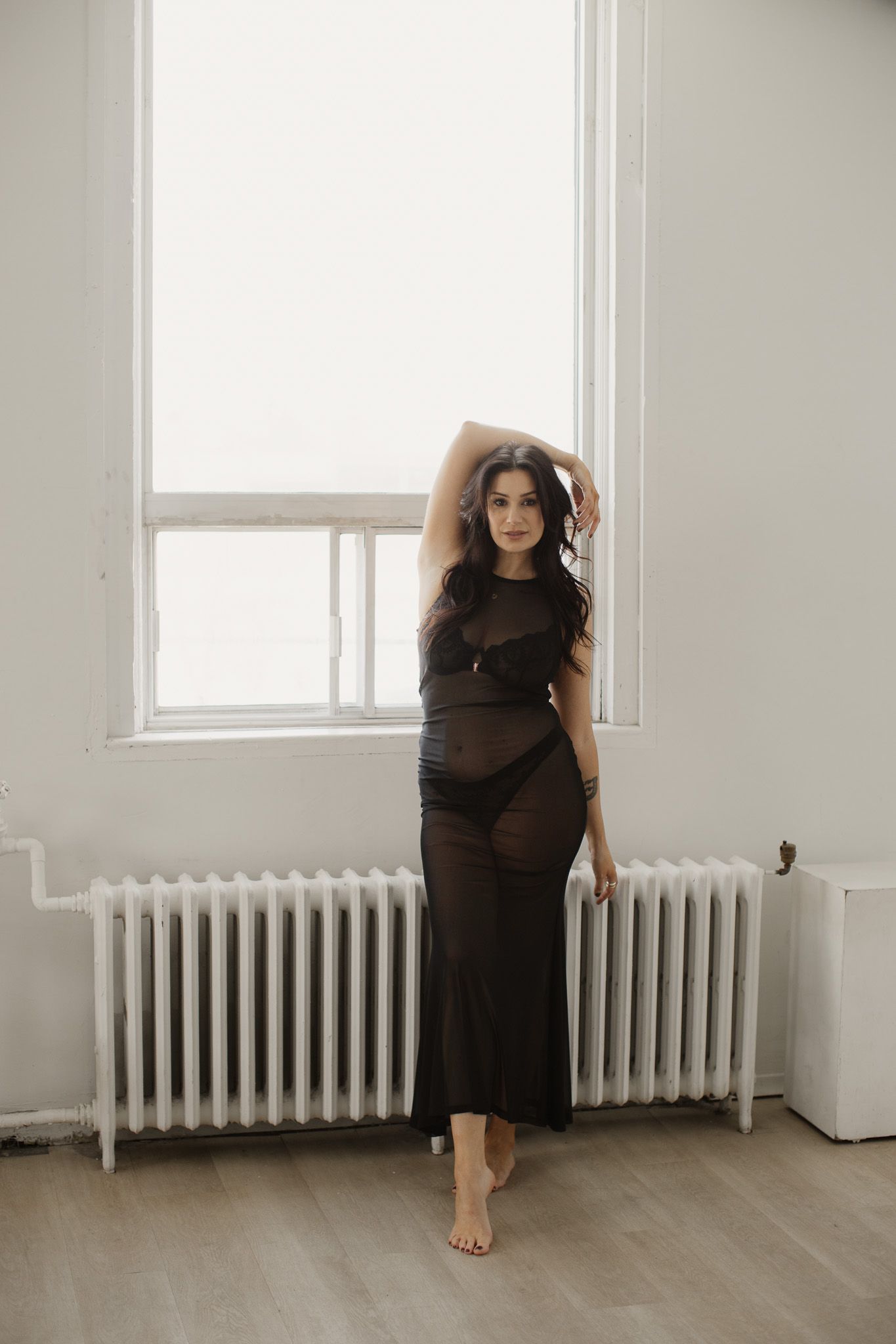 Person wearing a long black dress poses barefoot against a white wall with a radiator and window in a minimalist setting.