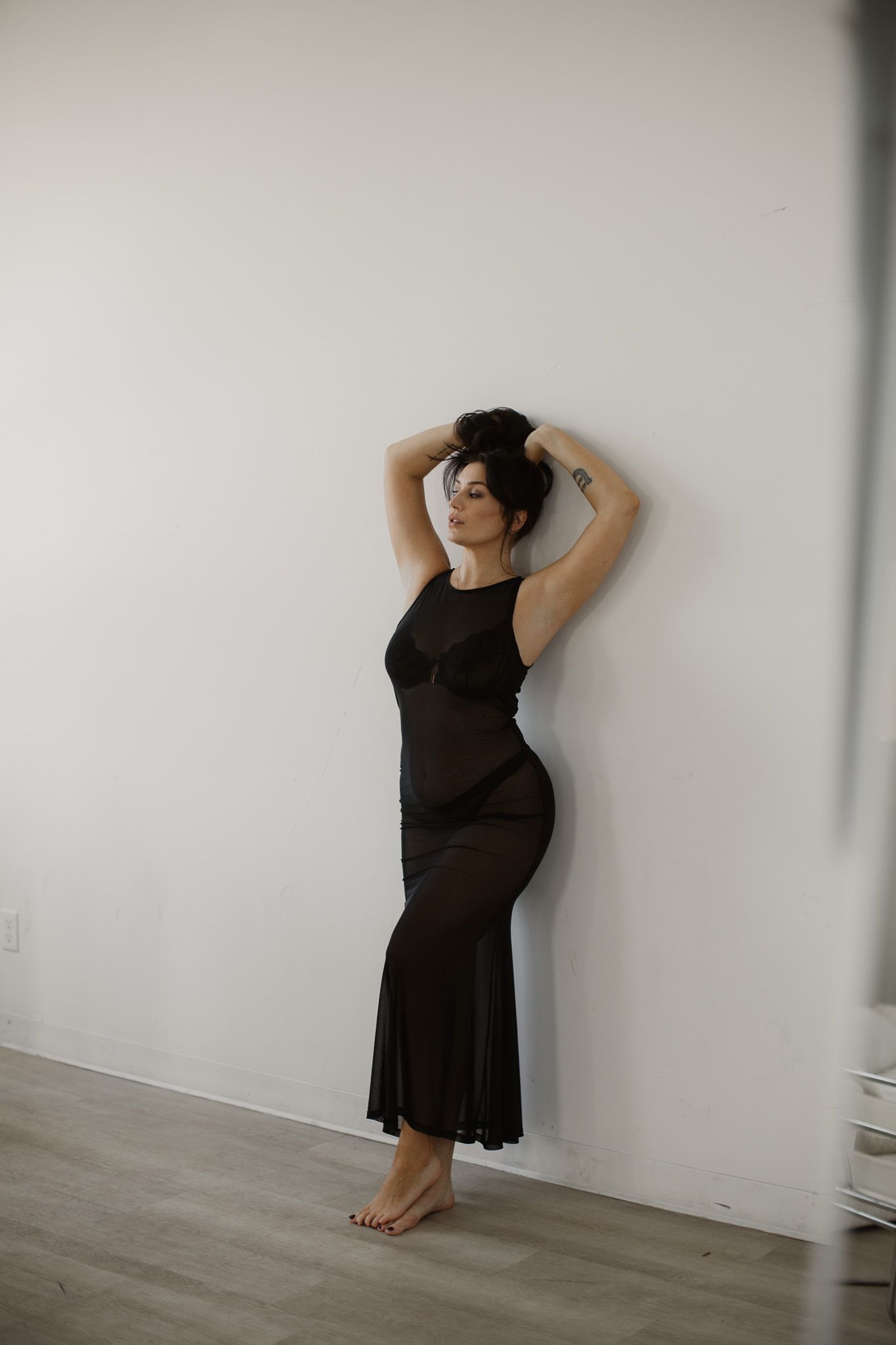 A person in a black dress poses elegantly against a white wall in a minimalist setting with hardwood floors.