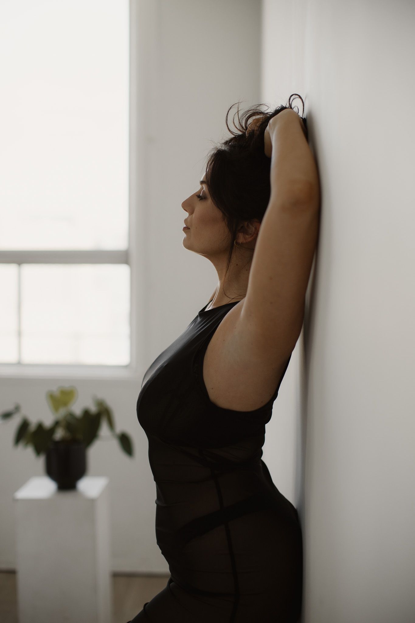 A silhouette of a person in a dark outfit stretches with raised arms against a window in a minimalist indoor setting.