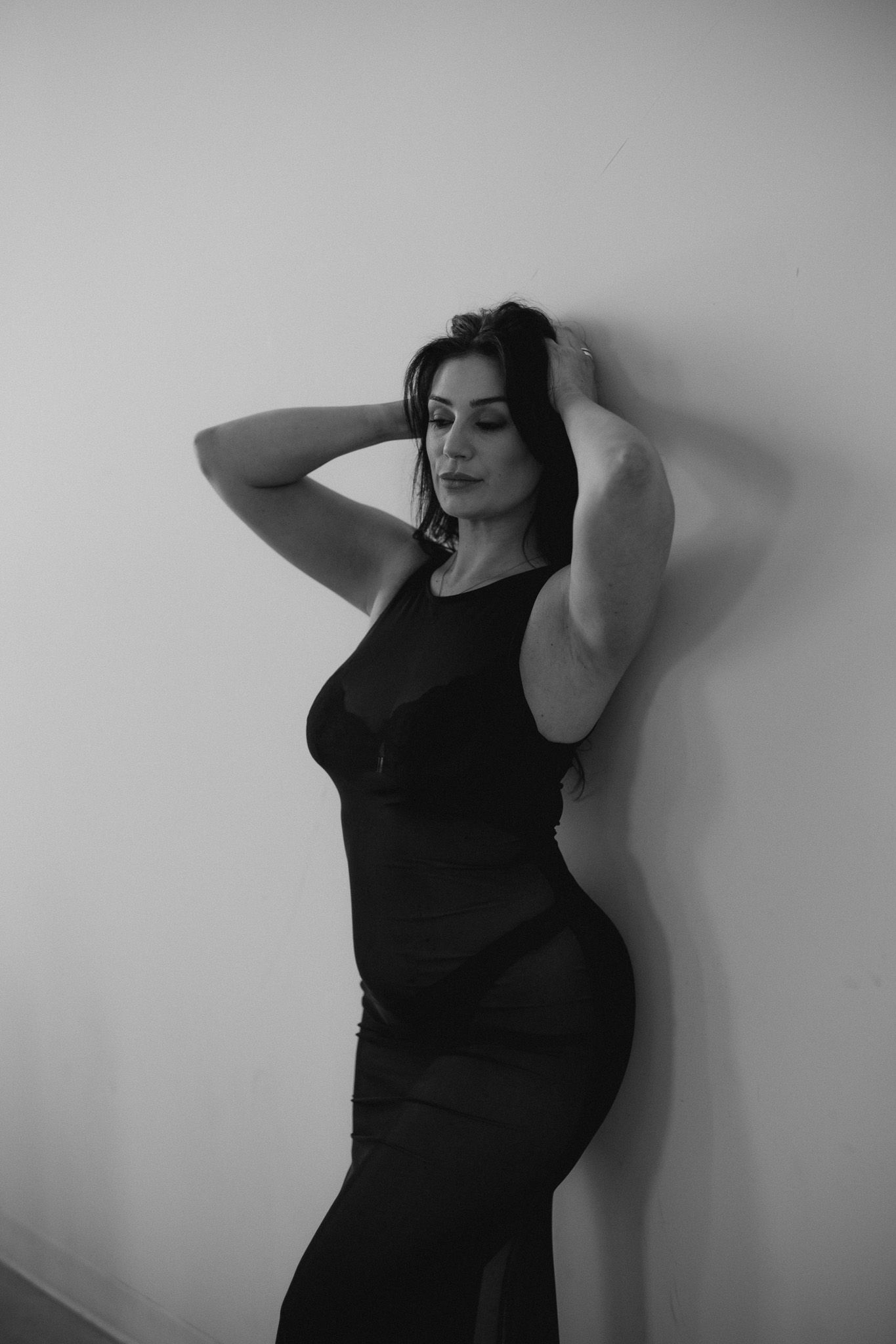 A person in a black fitted dress poses against a white wall in dramatic black and white photography.