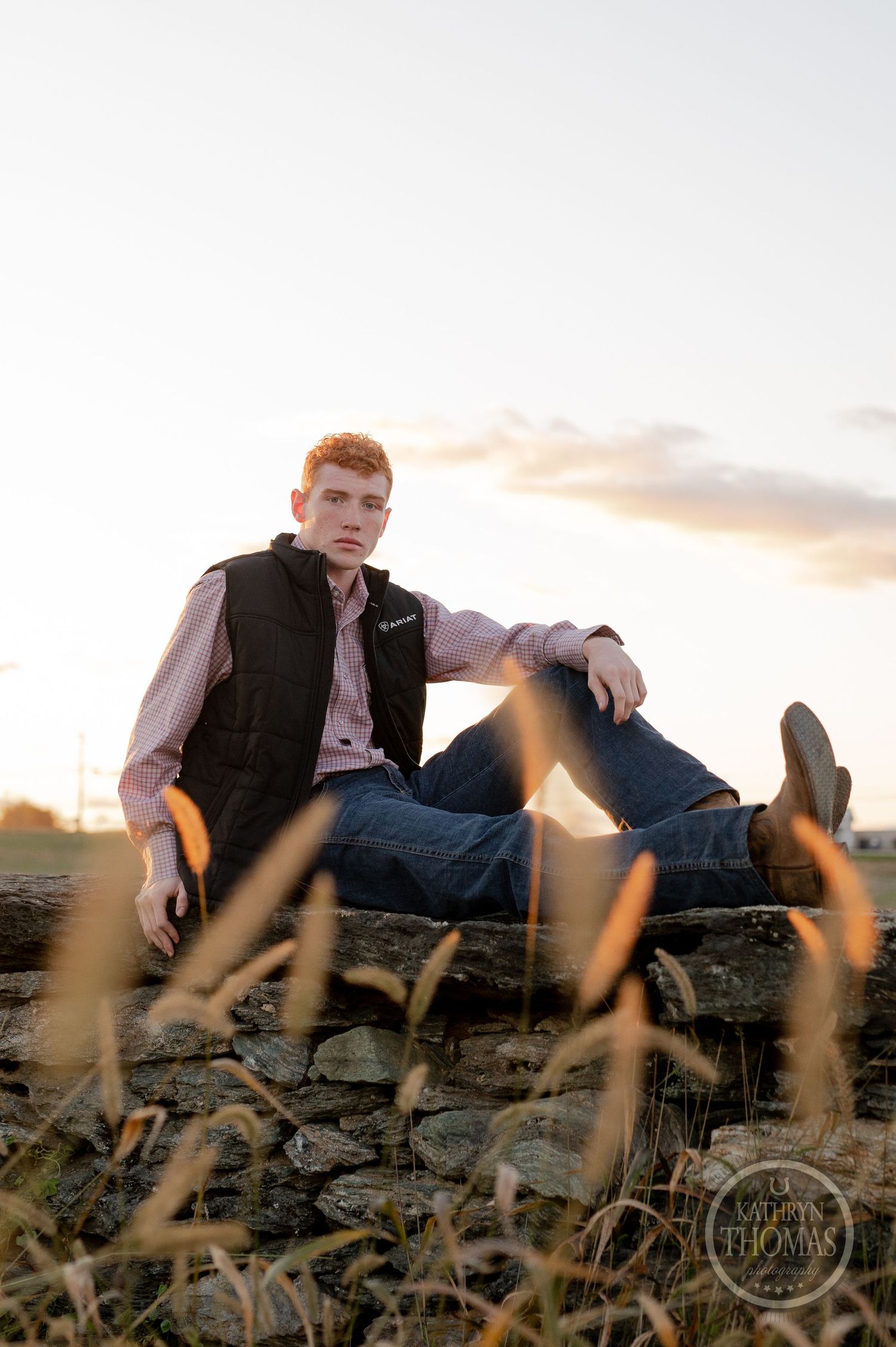 Person in vest and jeans relaxing on stone wall during sunset with tall grass below.