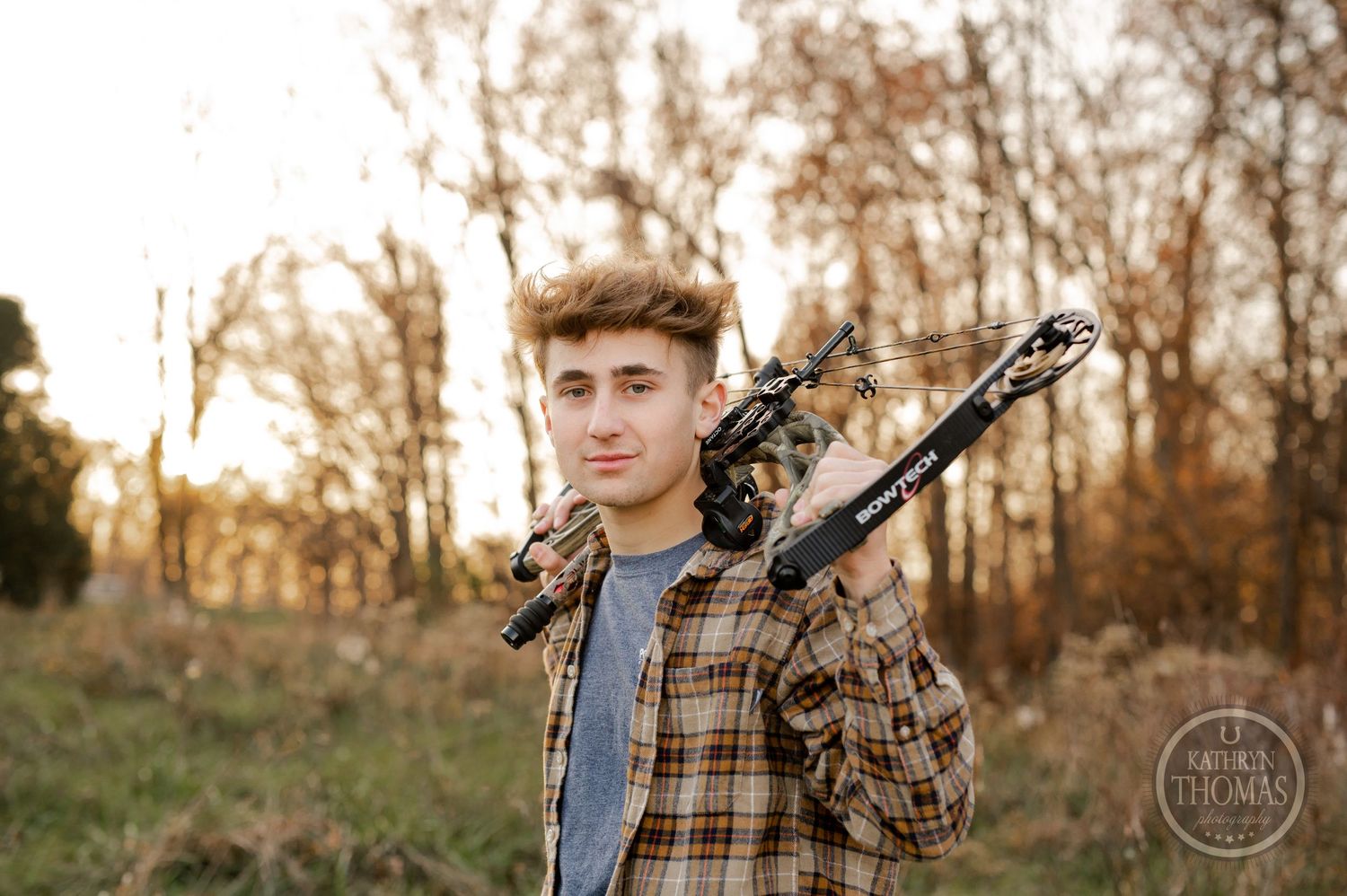 A person in a plaid flannel shirt carries archery equipment while standing in an autumn field at golden hour.