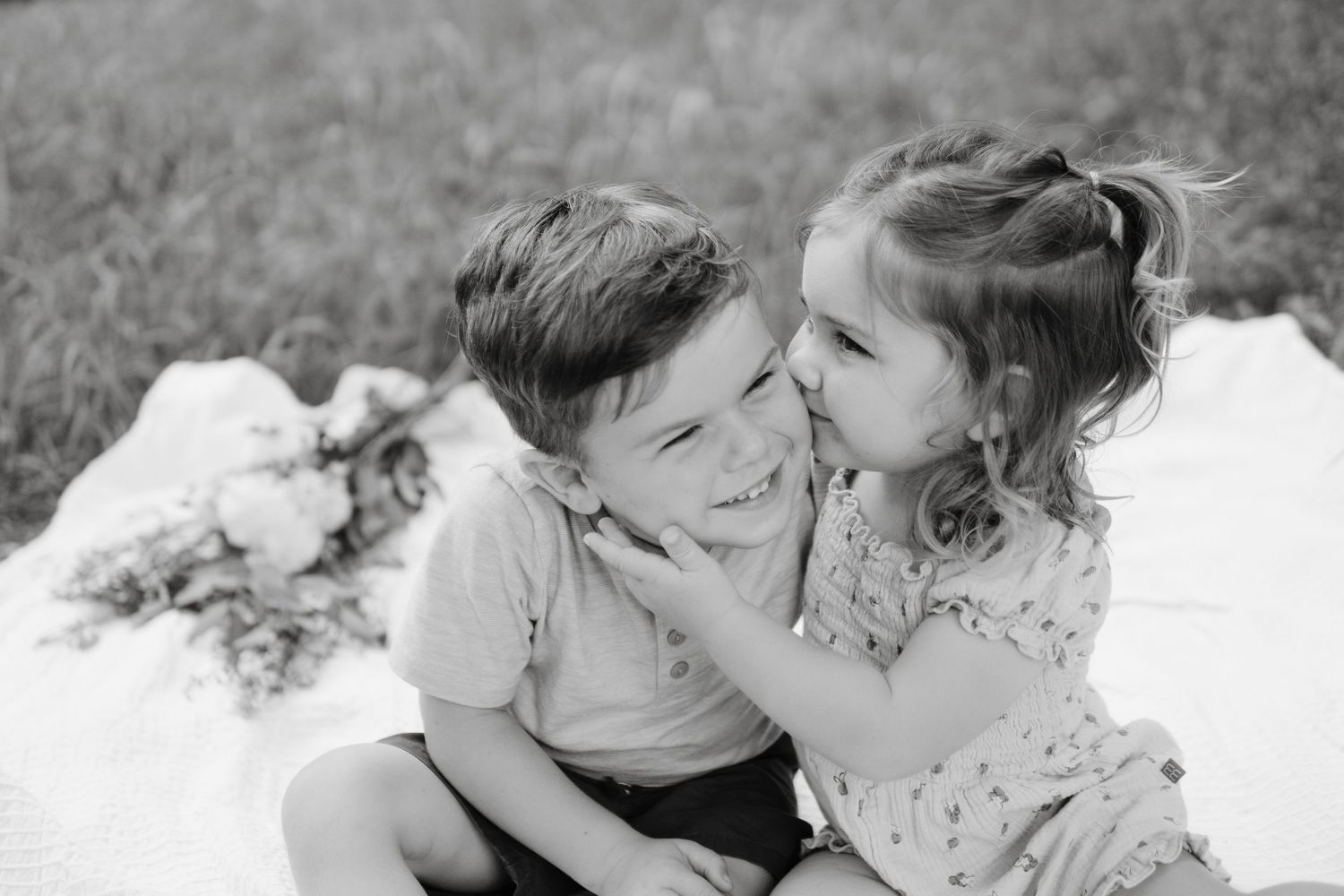 Black and white photograph showing two young children sitting close together outdoors and sharing a playful moment.