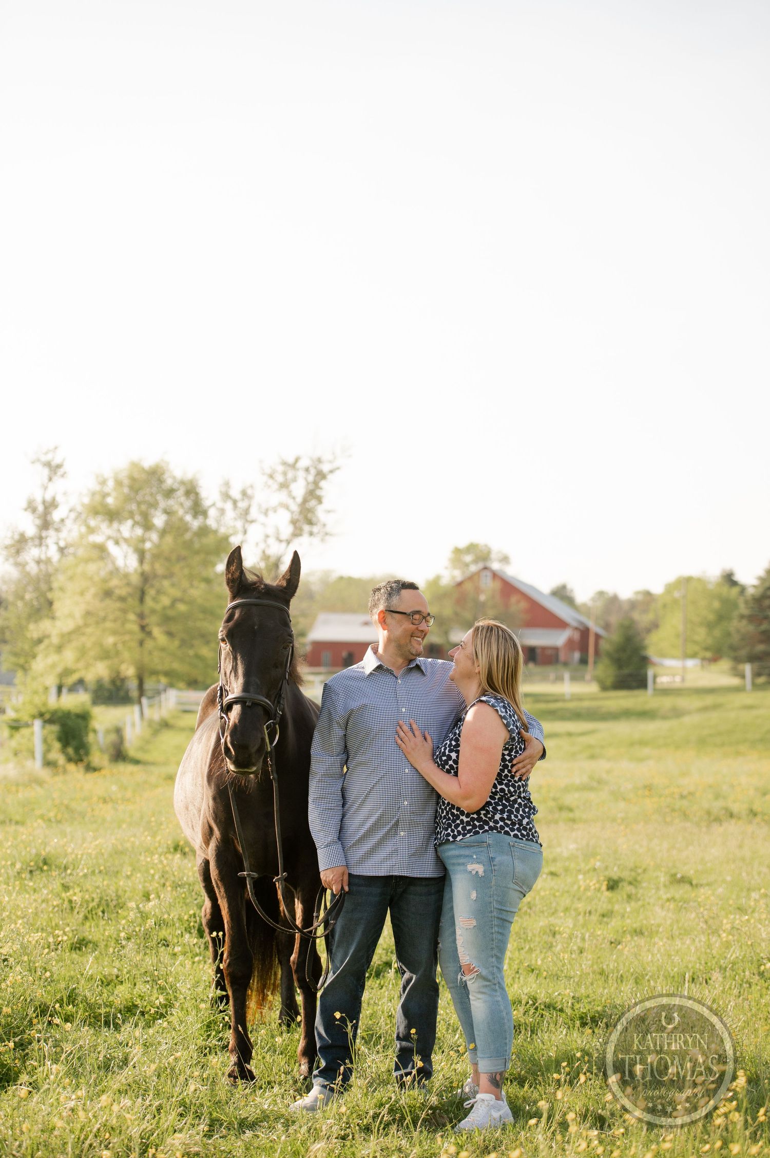 A couple stands close together with their horse in a sunlit field with a red barn in the distance.