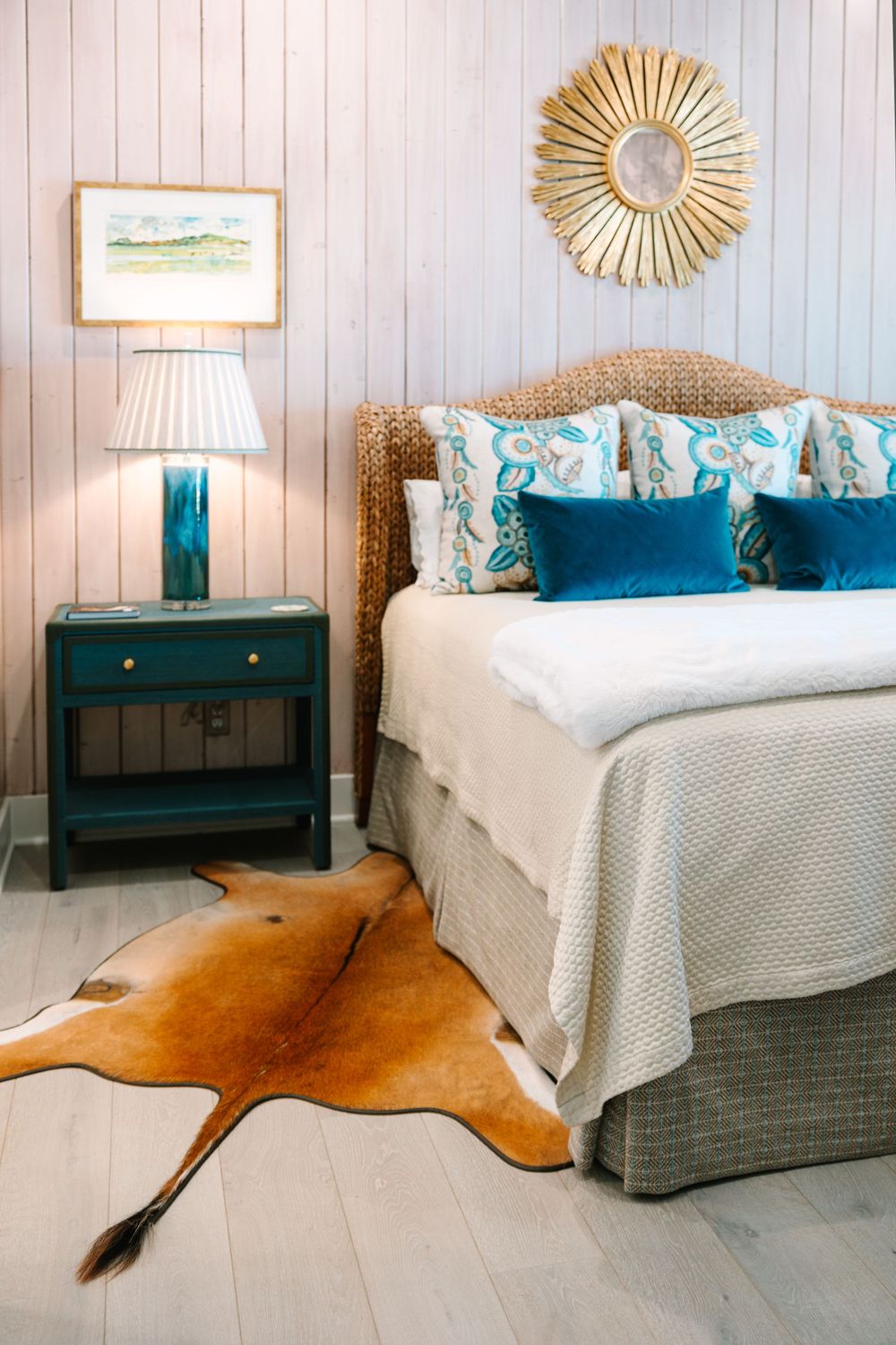 Coastal-themed bedroom with seagrass headboard, blue accents, and sunburst mirror above the bed.
