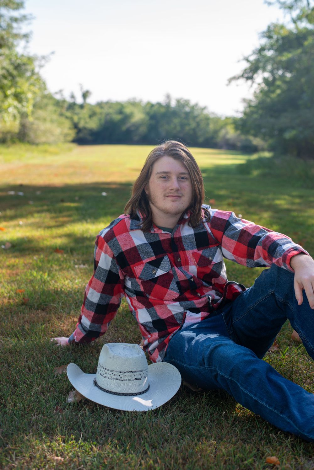 Person in red plaid shirt and jeans relaxes on grass with cowboy hat beside them.