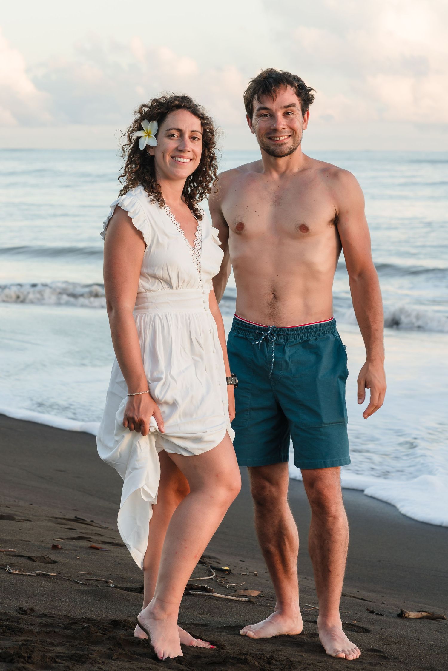 Couple poses together on black sand beach at sunset wearing casual beach attire.